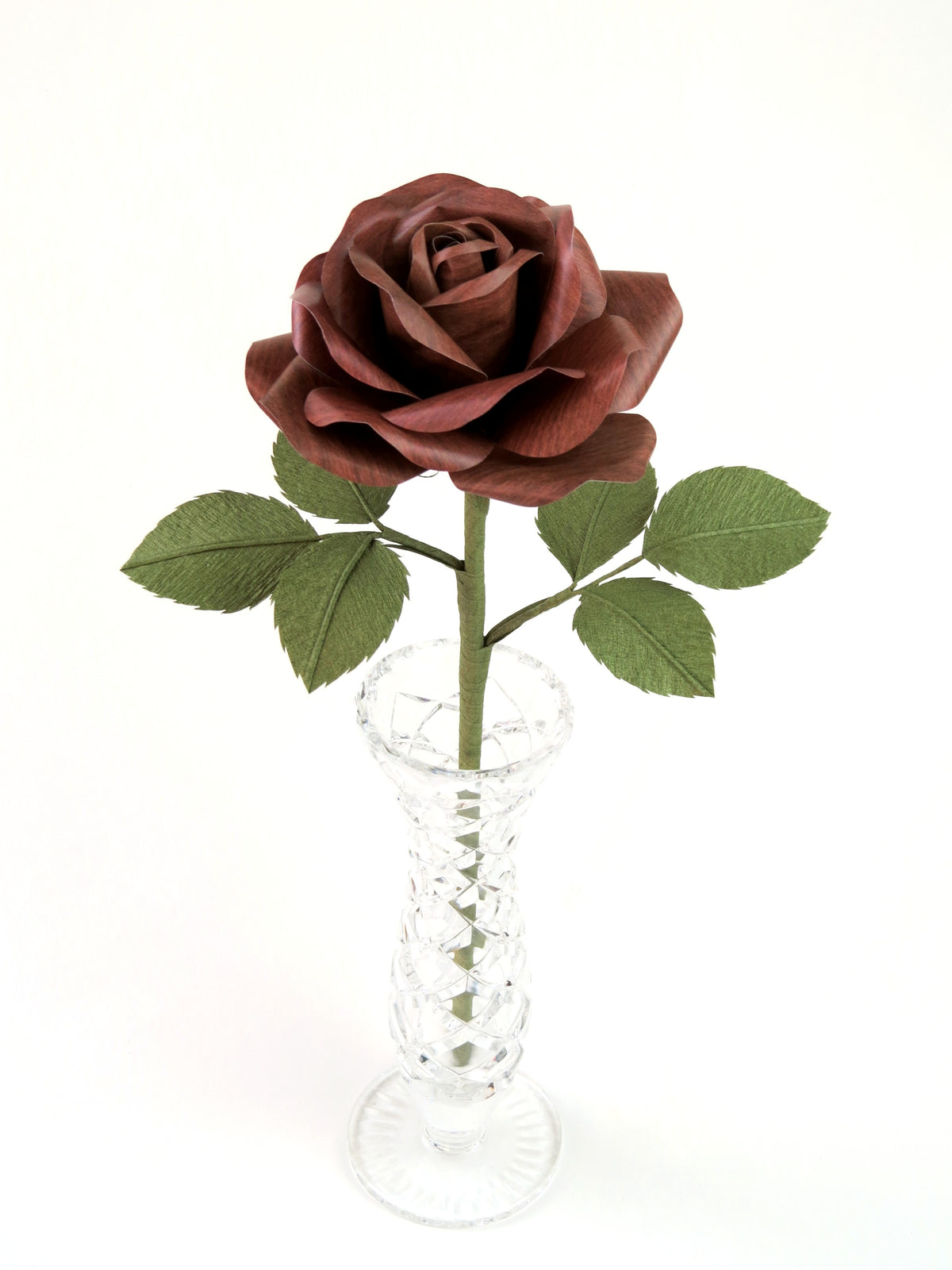 Dark wood grain paper rose with six green leaves standing in a narrow glass vase set against a white background