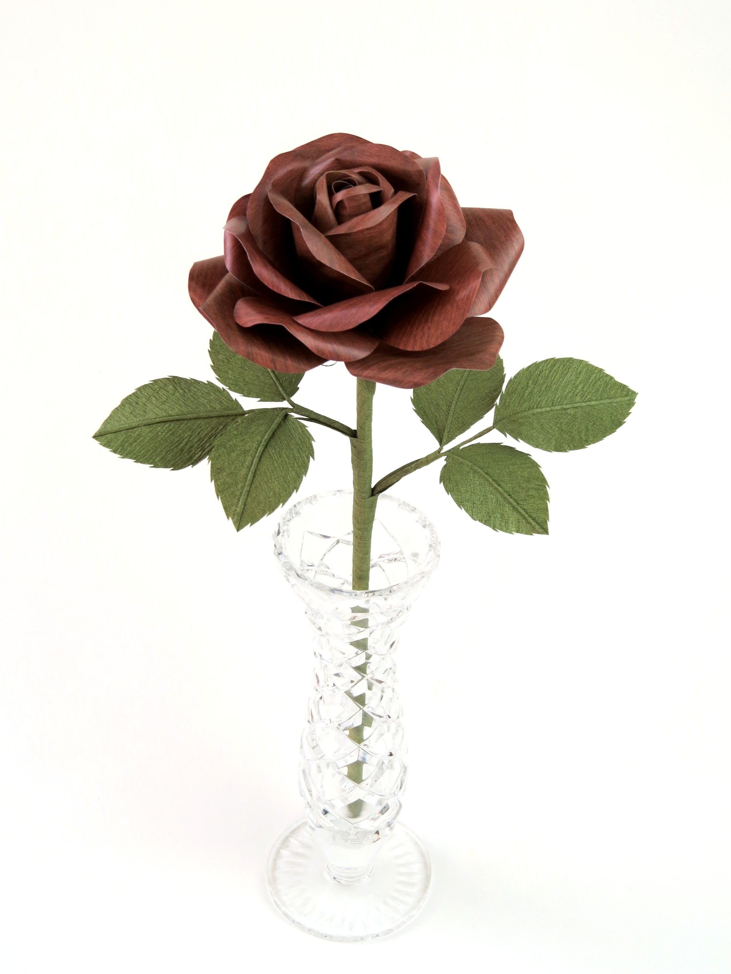 Dark wood grain paper rose with six green leaves standing in a narrow glass vase set against a white background