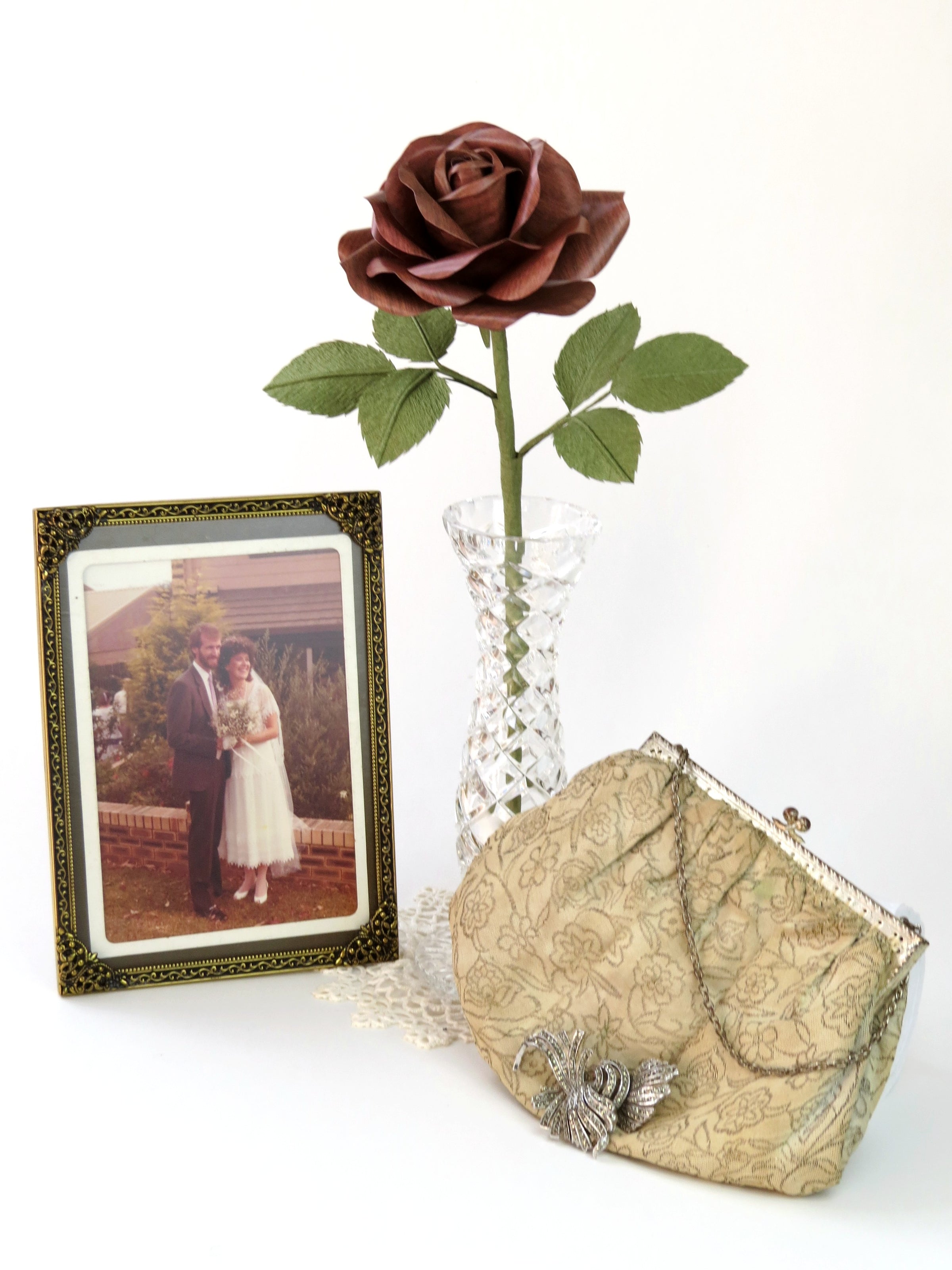 Dark wood grain paper rose with six green leaves standing in a slender glass vase on top of a white doily with a framed vintage wedding photo of a happy bride and groom standing beside it. To the right of the vase is a cream and silver floral vintage purse with a marcasite brooch resting on the front of it. 