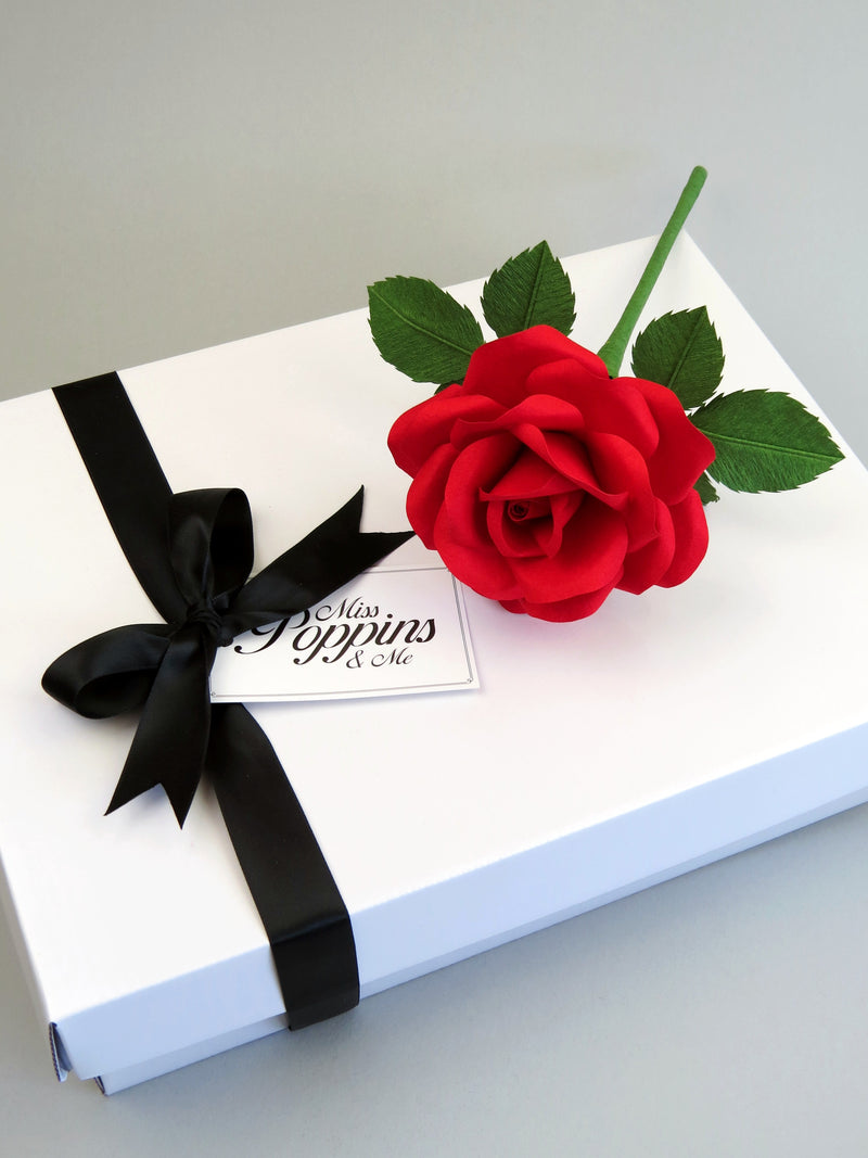Red cotton paper rose lying on top of a luxury white gift box that has a black satin ribbon tied in a bow with a Miss Poppins and Me gift tag attached