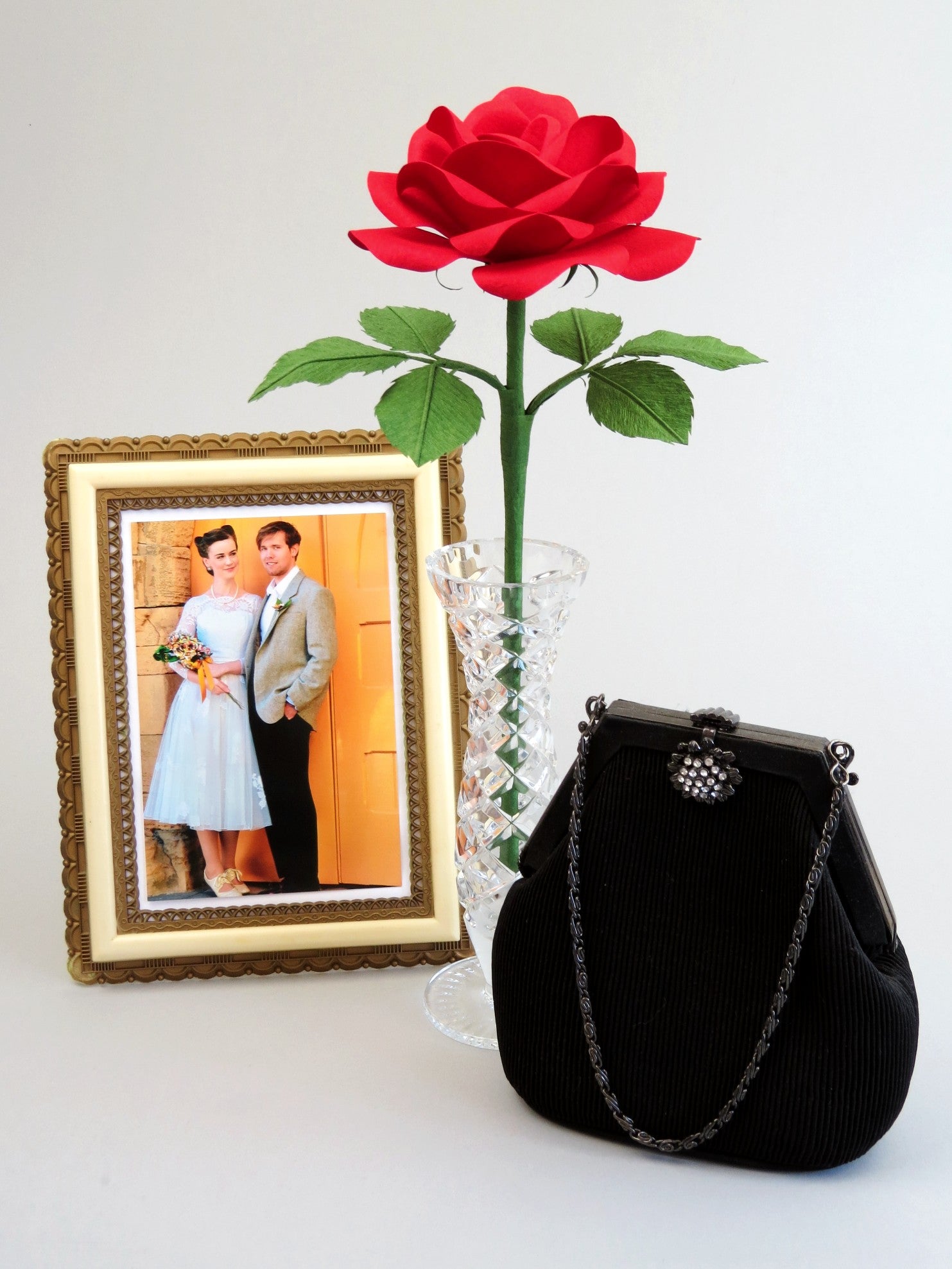 Red cotton paper rose with six green leaves standing in a slender glass vase with a framed wedding photo of a happy bride and groom to the left and a black vintage handbag with a diamante clutch and handing chain handle to the right of the vase