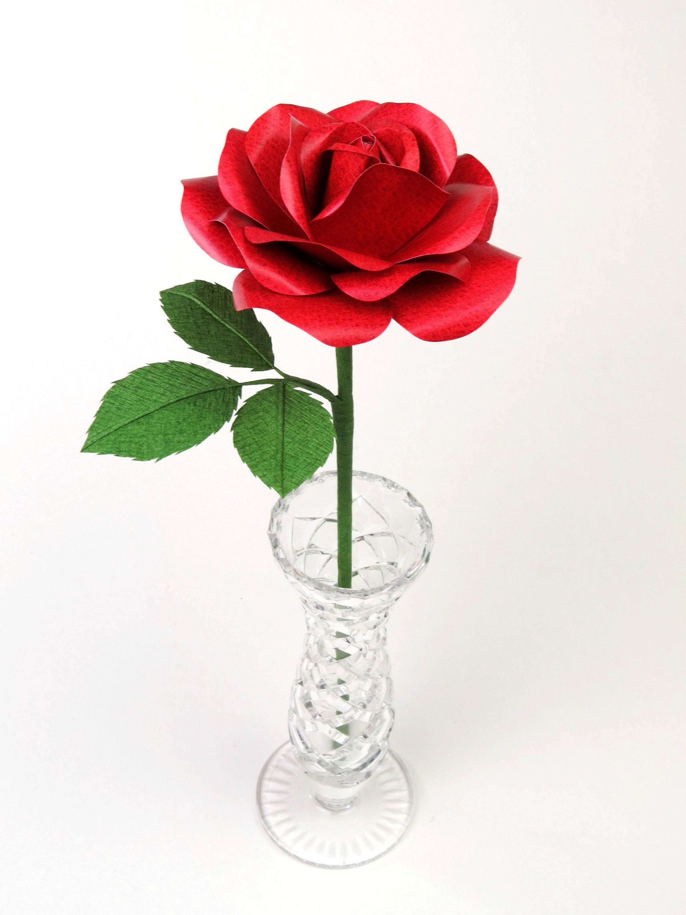 Red leather grain paper rose with three leaves standing in a narrow glass vase against a grey backdrop