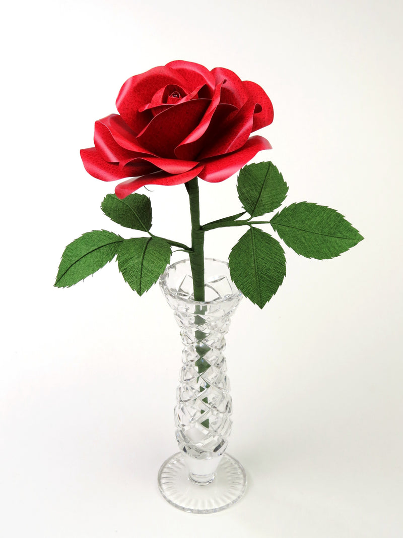 Red leather grain paper rose with six green leaves standing in a narrow glass vase set against a grey background