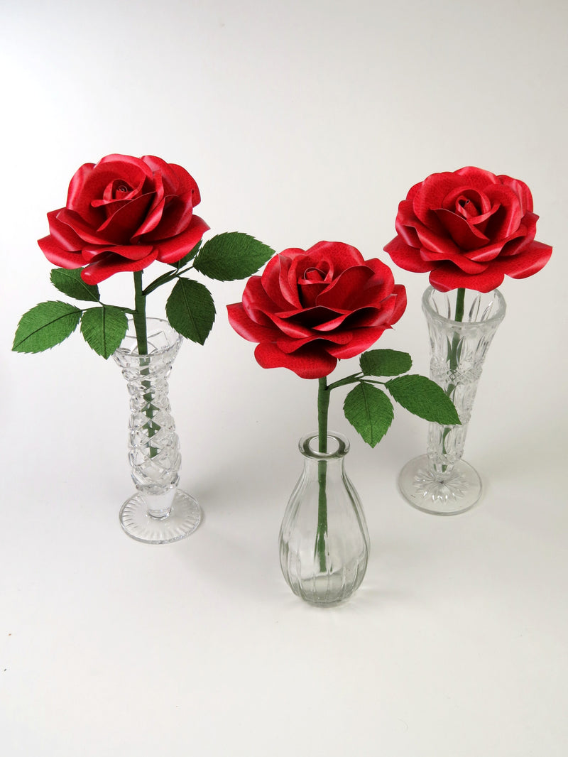 Three red leather grain paper roses standing upright in three different glass vases. The left rose has six green leaves attached, the middle rose has three leaves and the right rose has no leaves.