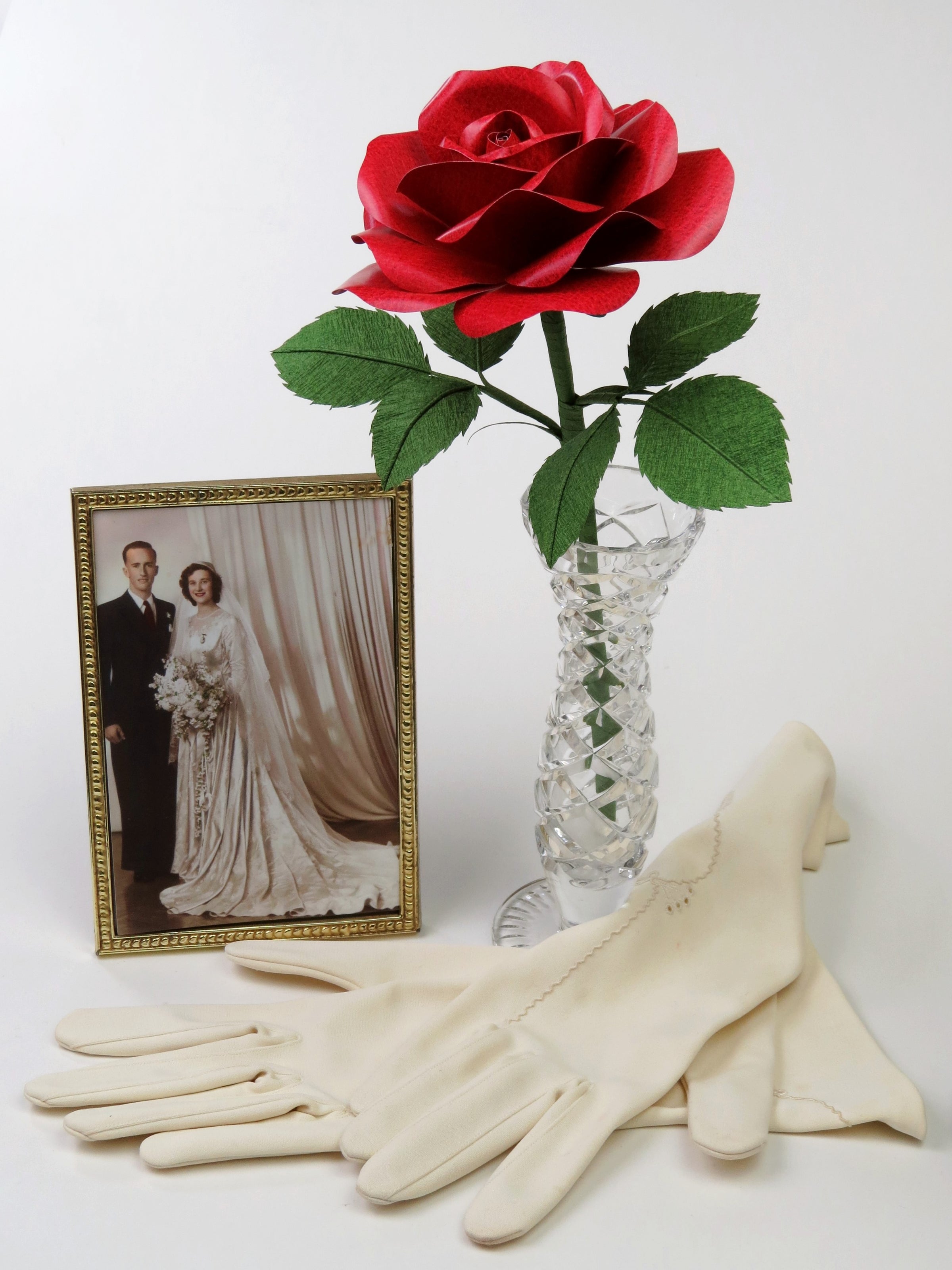 Red leather grain paper rose with six green leaves standing in a slender glass vase with a framed wedding photo of a happy bride and groom standing beside it. Lying at the front is a pair of vintage cream gloves. 