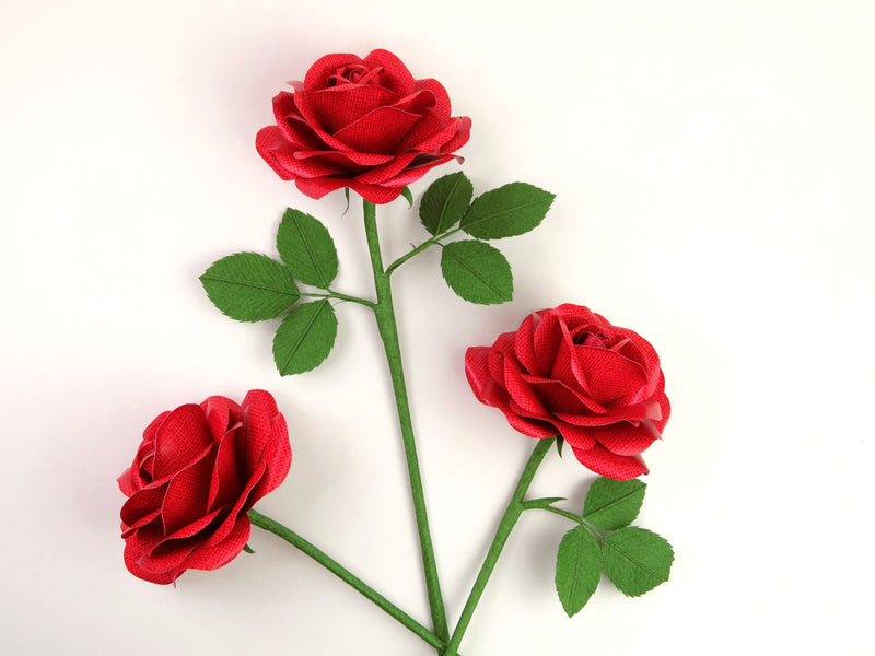 Three red linen grain paper roses randomly lying next to each other on a light grey background. The left rose has no leaves, the middle rose has six green leaves and the right rose has three leaves.