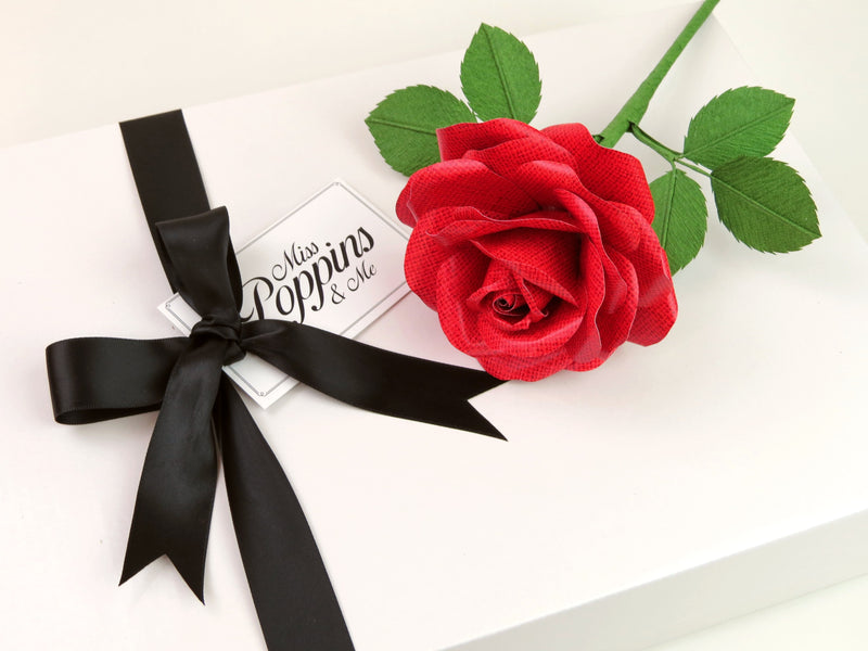 Detailed close up of a red linen grain paper rose lying on top of a luxury white gift box that has a black satin ribbon tied in a bow with a Miss Poppins and Me gift tag attached