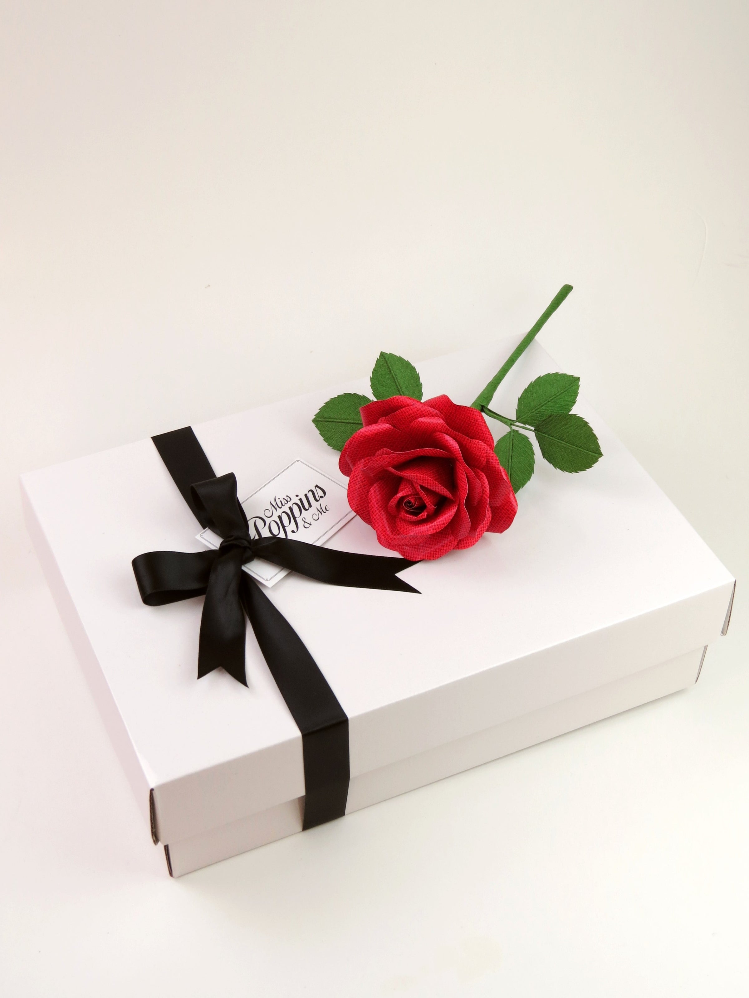 Red linen grain paper rose lying on top of a luxury white gift box that has a black satin ribbon tied in a bow with a Miss Poppins and Me gift tag attached