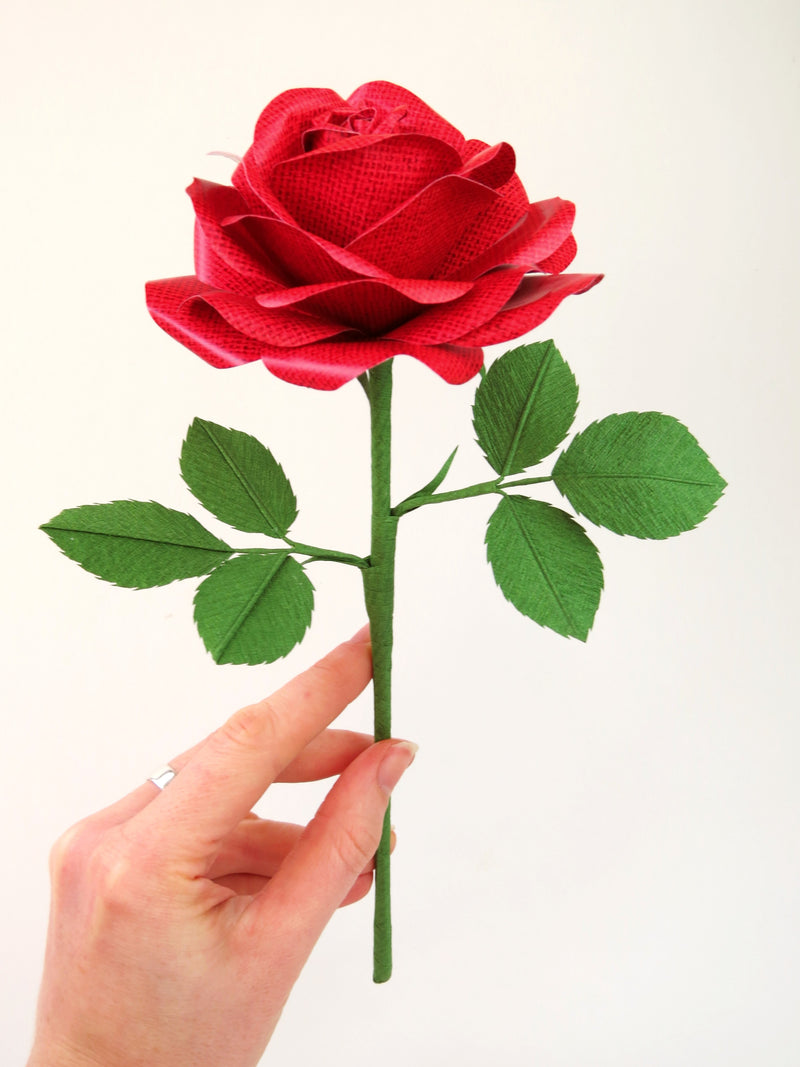 Pale white hand delicately holding the stem of a red linen grain paper rose with six leaves