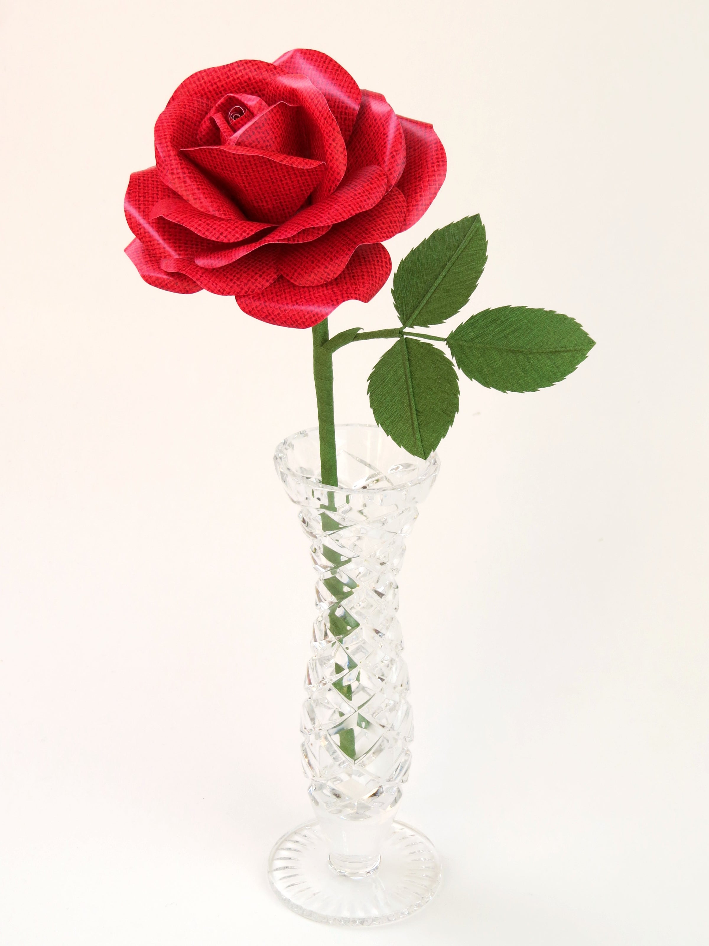 Red linen grain paper rose with three leaves standing in a narrow glass vase against a light grey backdrop