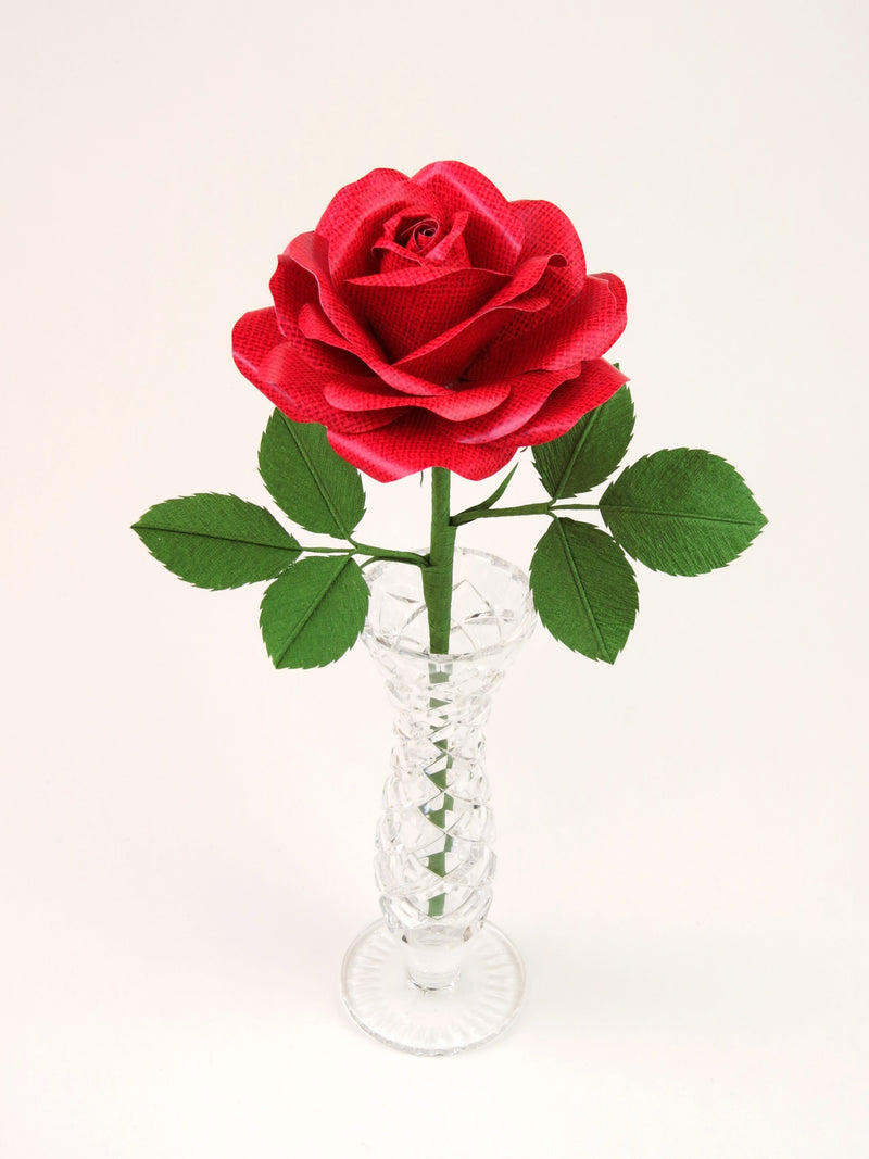 Red linen grain paper rose with six green leaves standing in a narrow glass vase set against a light grey background