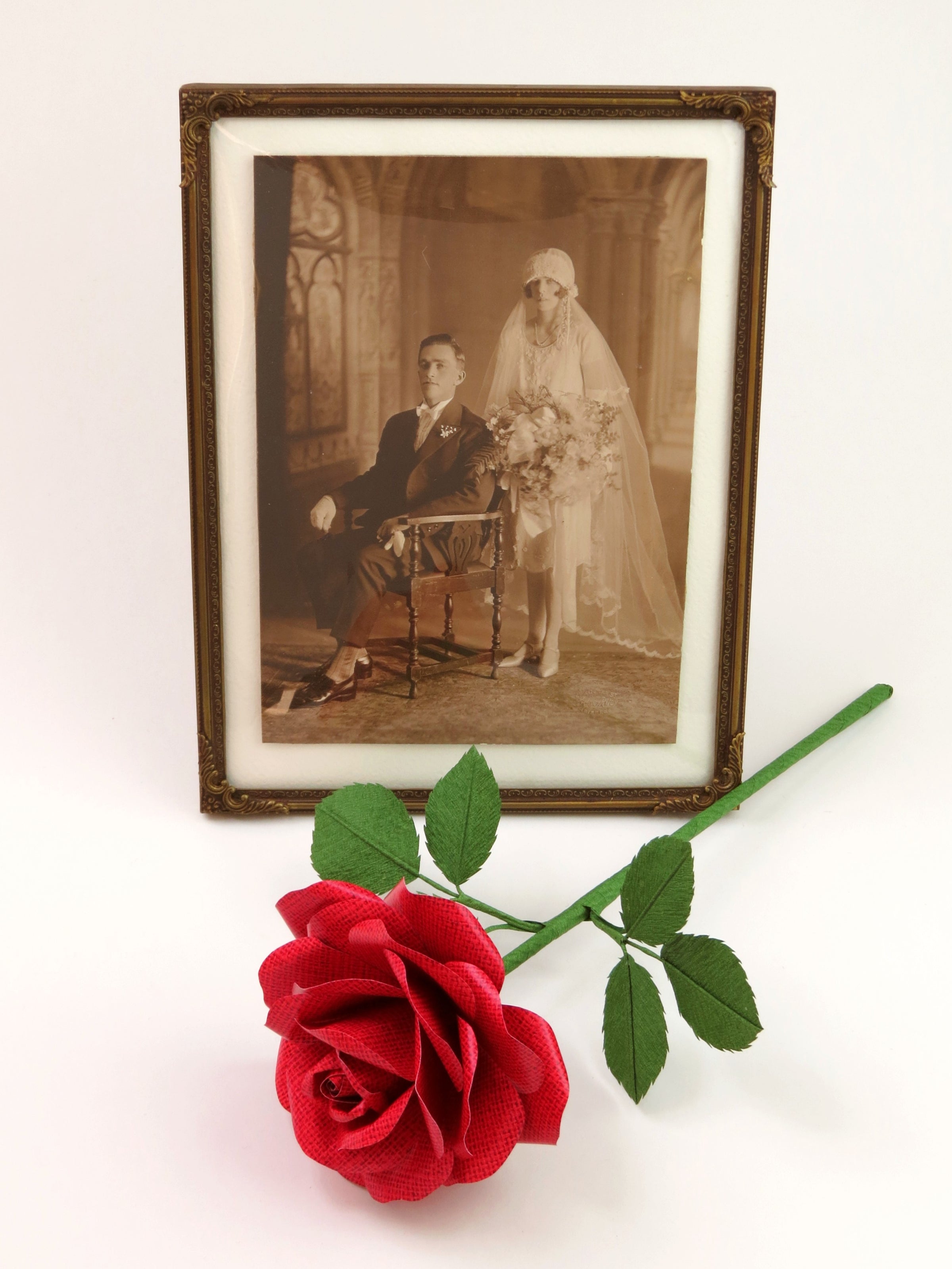 Red linen grain paper rose with six green leaves lying in front of a framed 1920’s wedding photo 