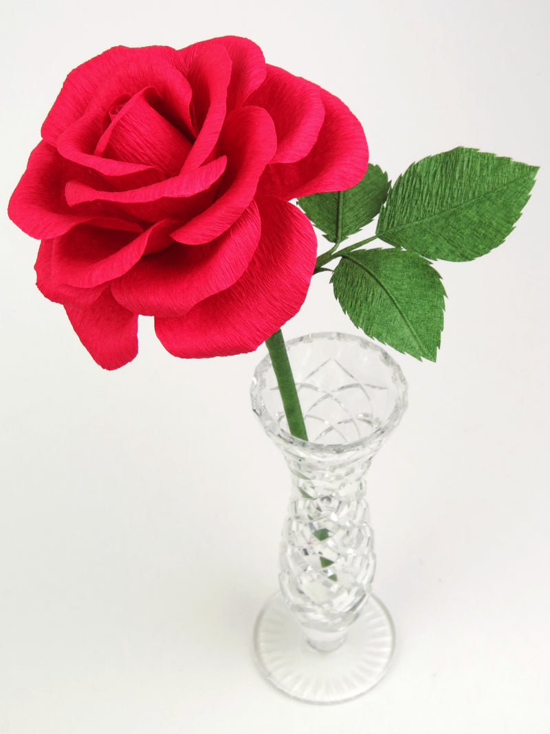 Red paper rose with three leaves standing in a narrow glass vase against a white backdrop