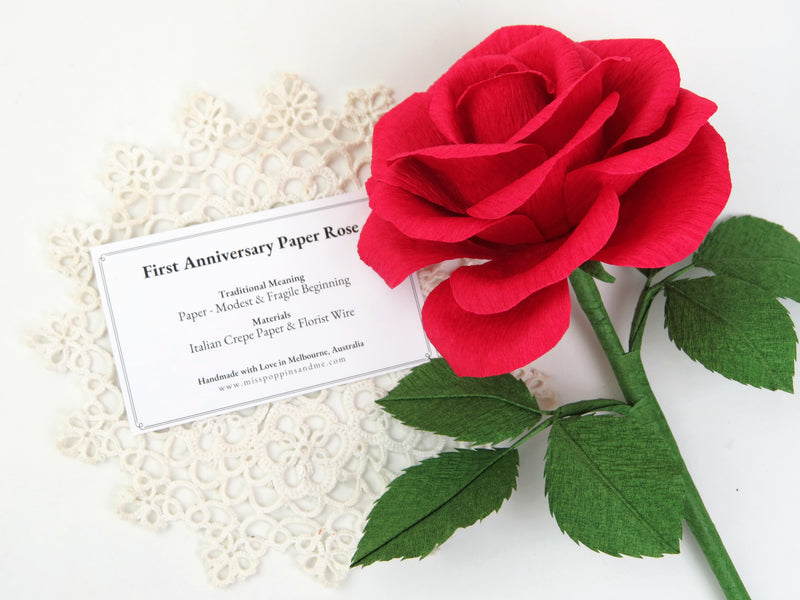 Red paper rose lying on a small white vintage doily which has a product tag resting on it detailing the meaning and materials of this paper rose