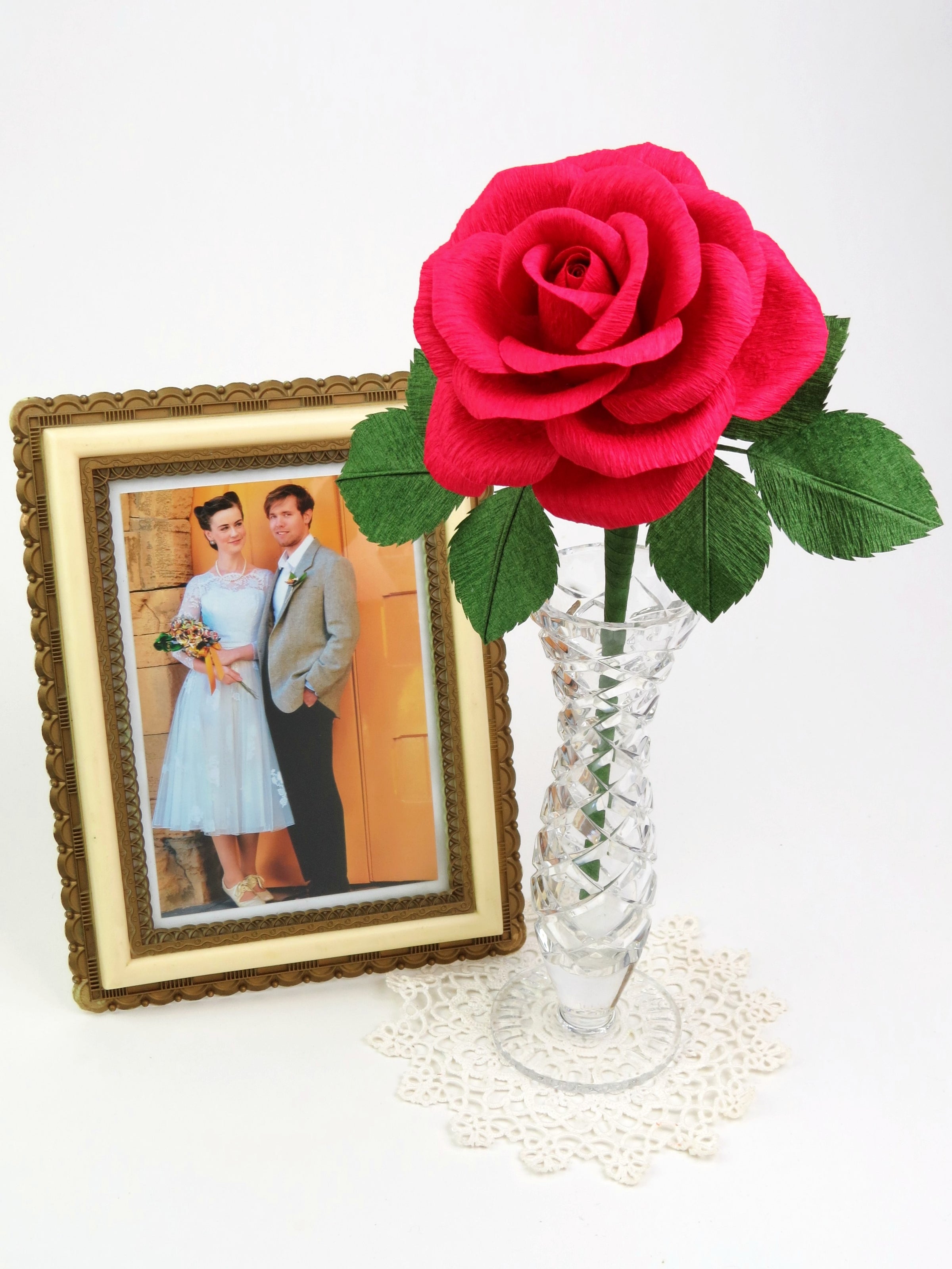 Red paper rose with six green leaves standing in a slender glass vase on top of a white vintage doily with a framed wedding photo of a happy bride and groom standing beside it