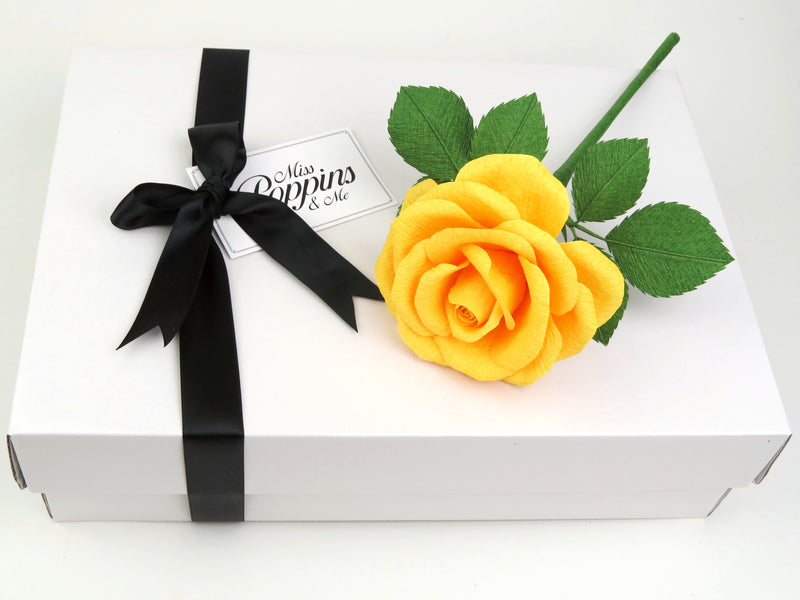 Yellow paper rose lying on top of a luxury white gift box that has a black satin ribbon tied in a bow with a Miss Poppins and Me gift tag attached