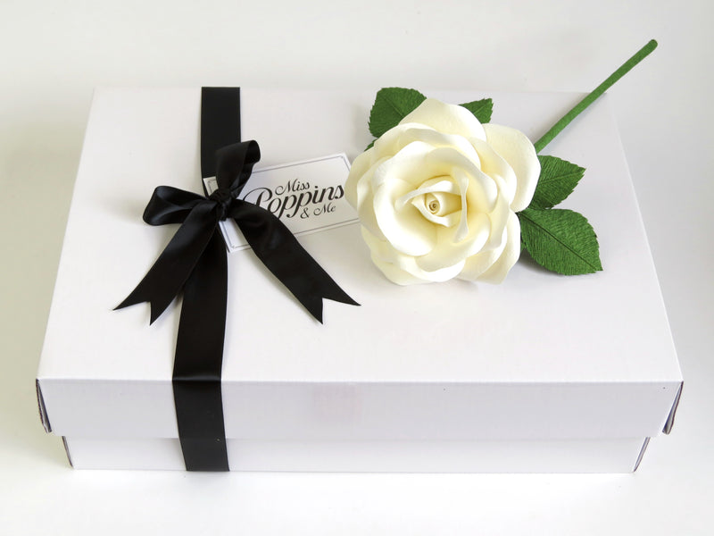 White cotton paper rose with green leaves lying on top of a luxury white gift box that has a black satin ribbon tied in a bow with a Miss Poppins and Me gift tag attached