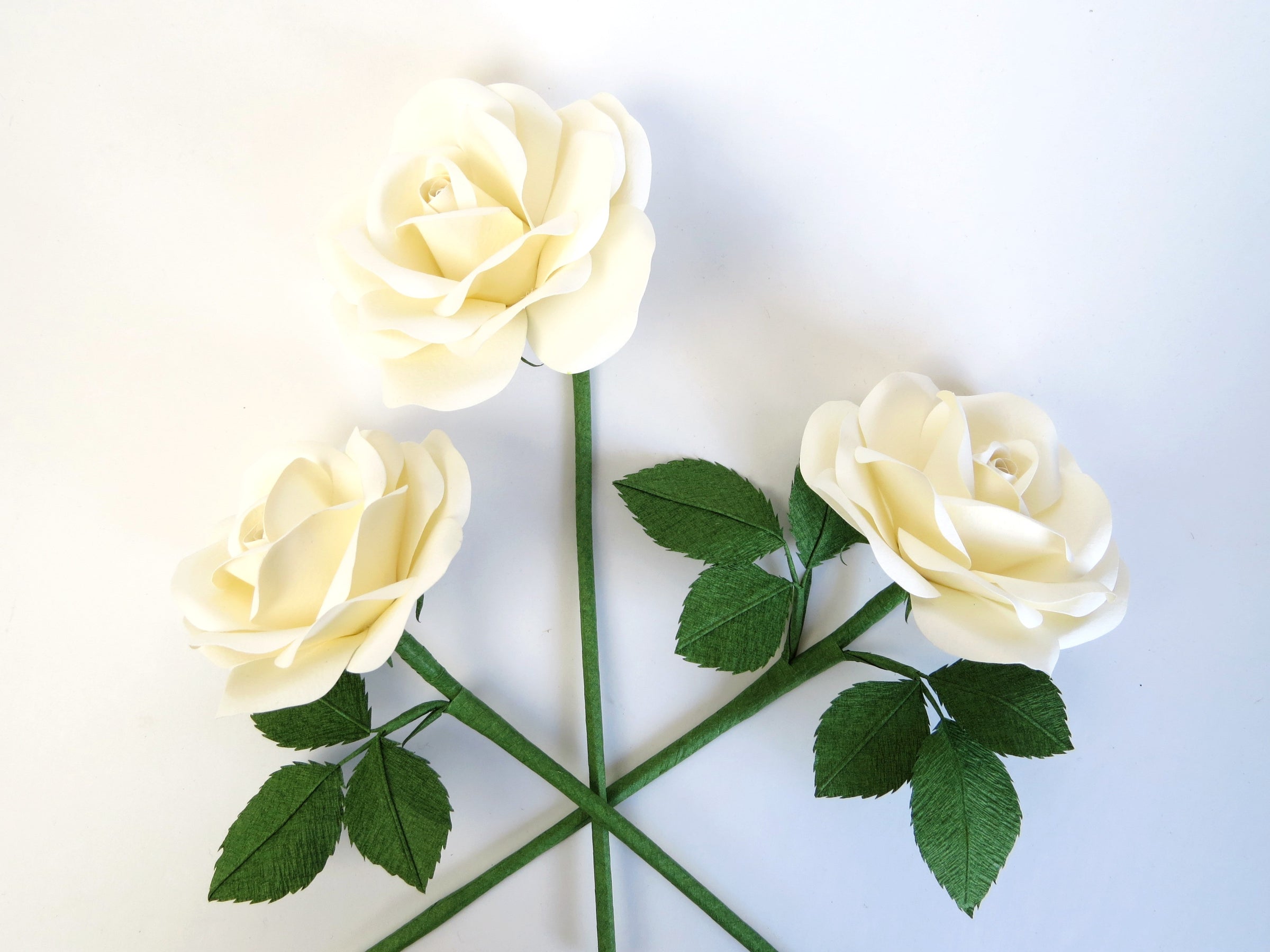 Three white cotton paper roses randomly laid out next to each other on a light grey background. The left rose has three green leaves attached, the high middle rose has no leaves and the right rose has six green leaves