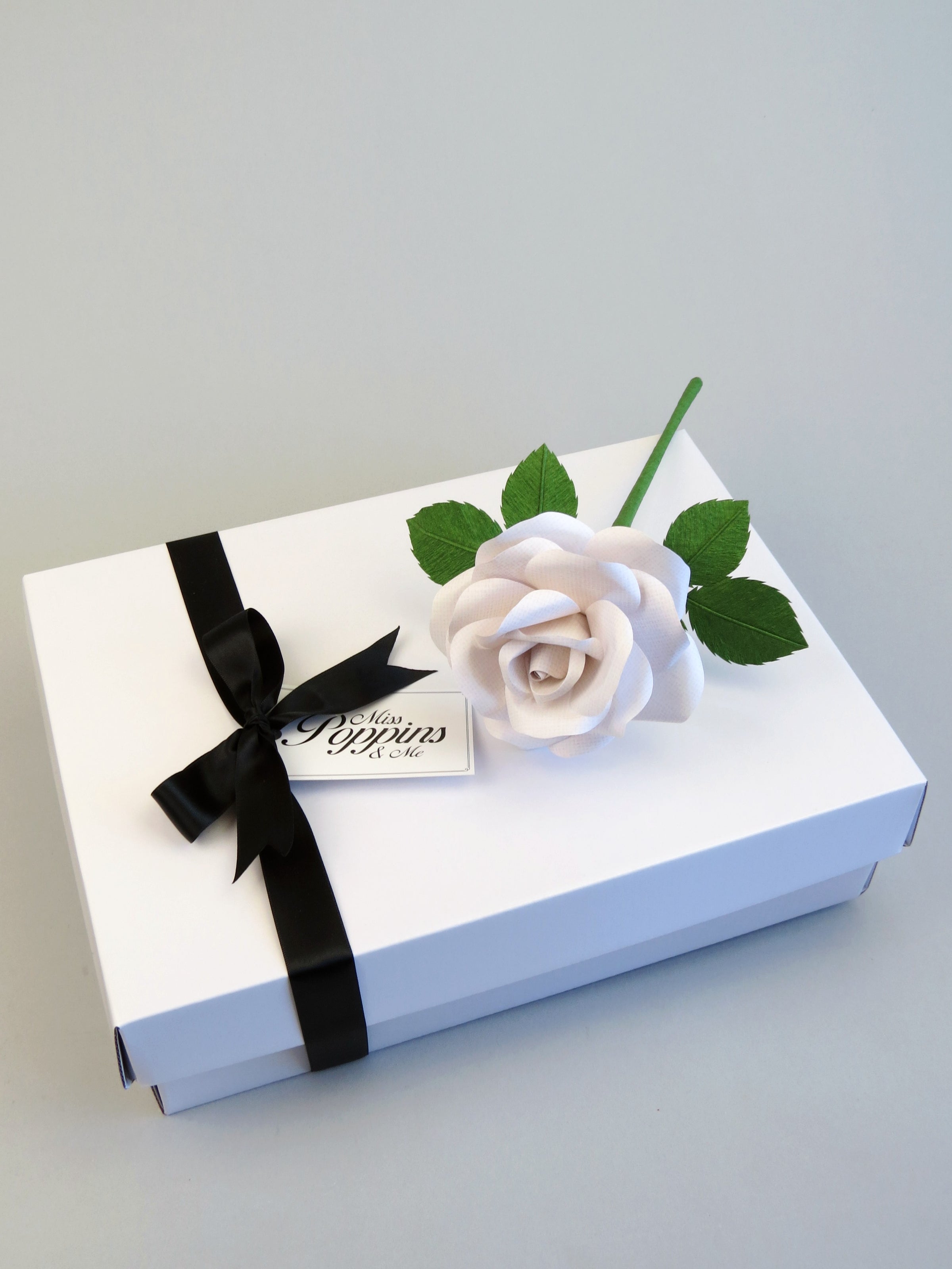 White linen grain paper rose lying on top of a luxury white gift box that has a black satin ribbon tied in a bow with a Miss Poppins and Me gift tag attached