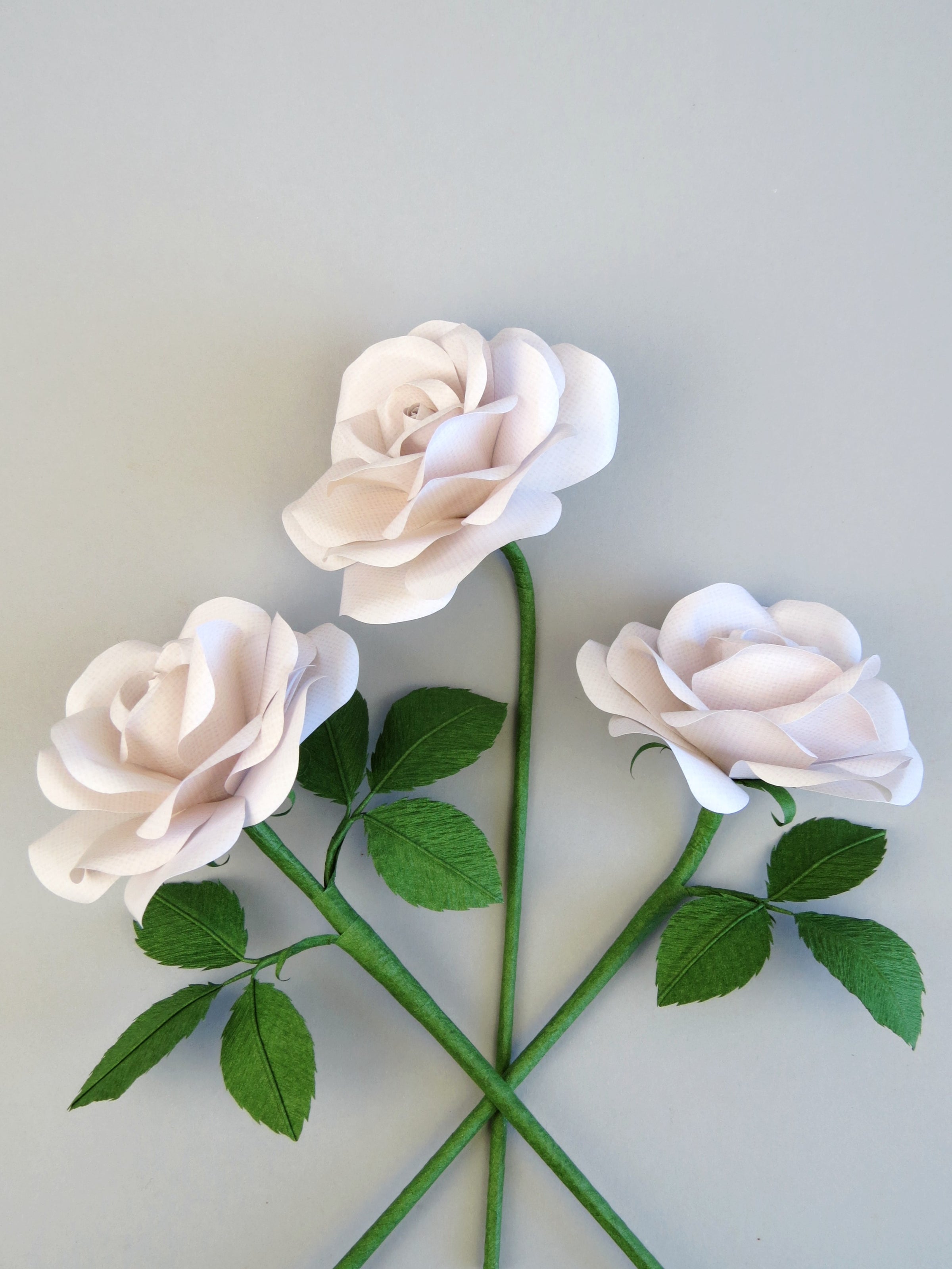 Three white linen grain paper roses randomly laid out next to each other on a light grey background. The left rose has six green leaves attached, the high middle rose has no leaves and the right rose has three leaves. 