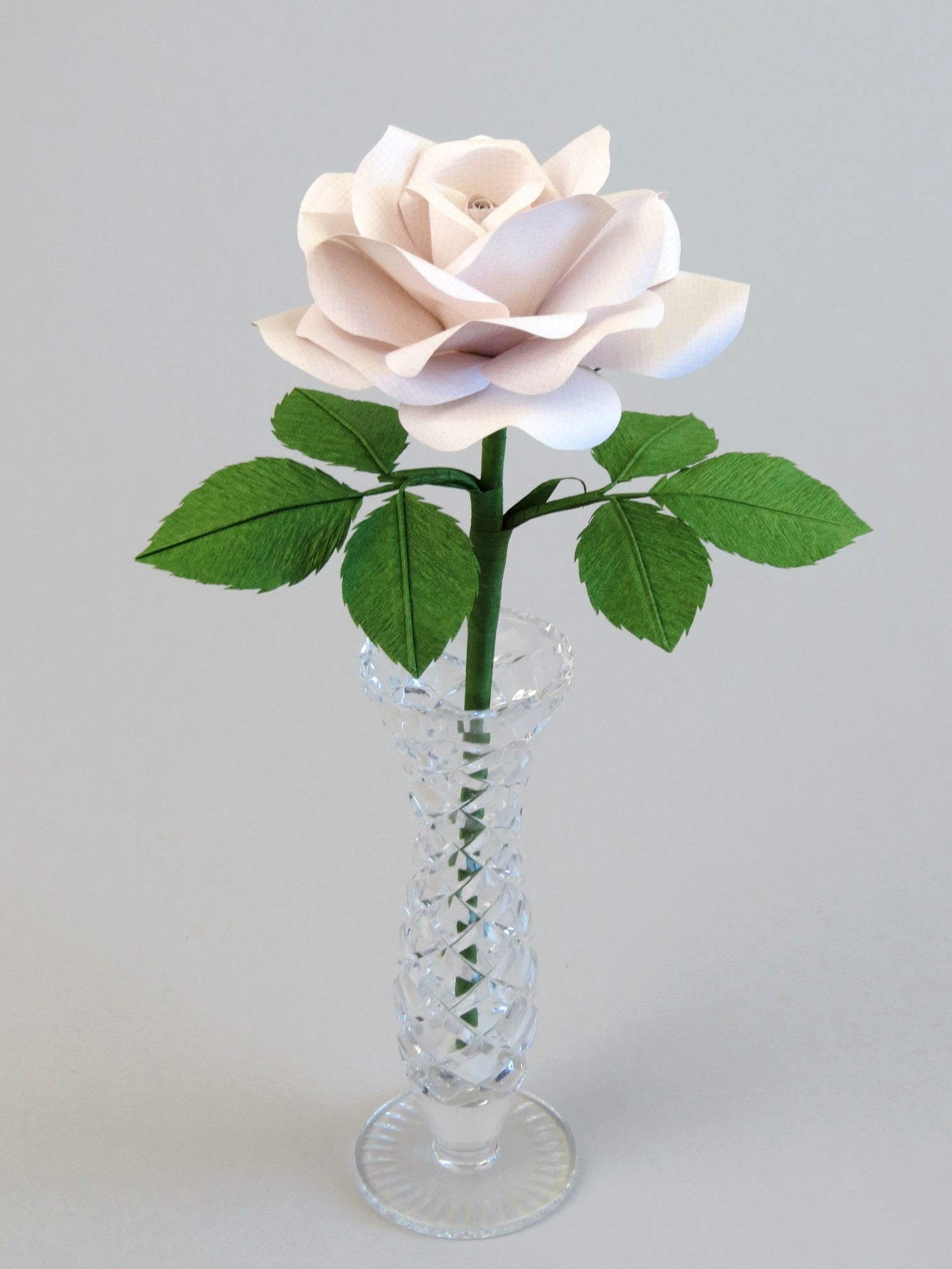 White linen grain paper rose with six leaves standing in a narrow glass vase against a light grey background