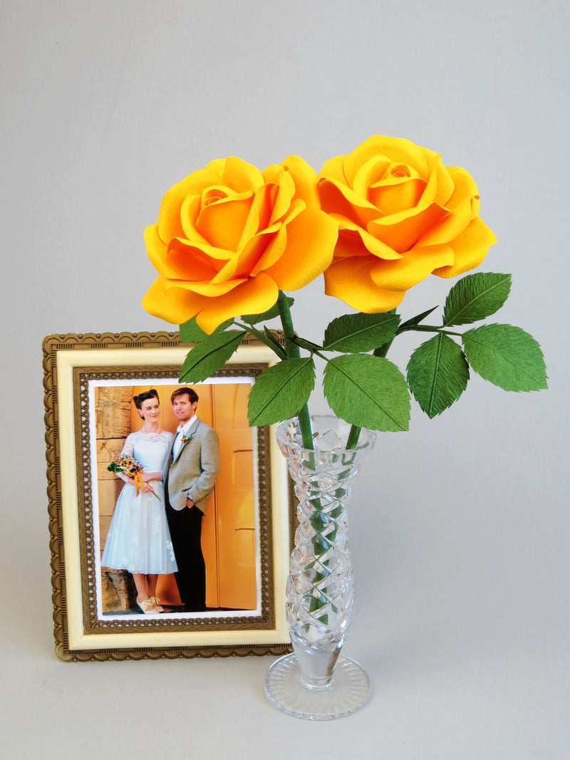 Yellow cotton paper rose bouquet of two roses with six green leaves each, perched in a slender glass vase with a framed vintage wedding photo of a happy couple standing beside it against a grey background