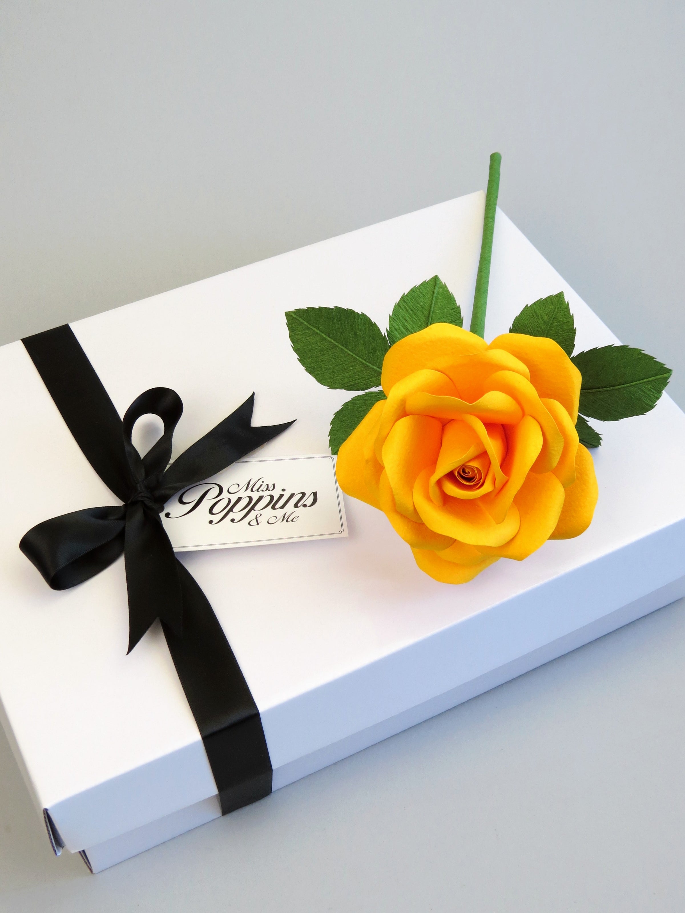 Yellow cotton paper rose lying on top of a luxury white gift box that has a black satin ribbon tied in a bow with a Miss Poppins and Me gift tag attached