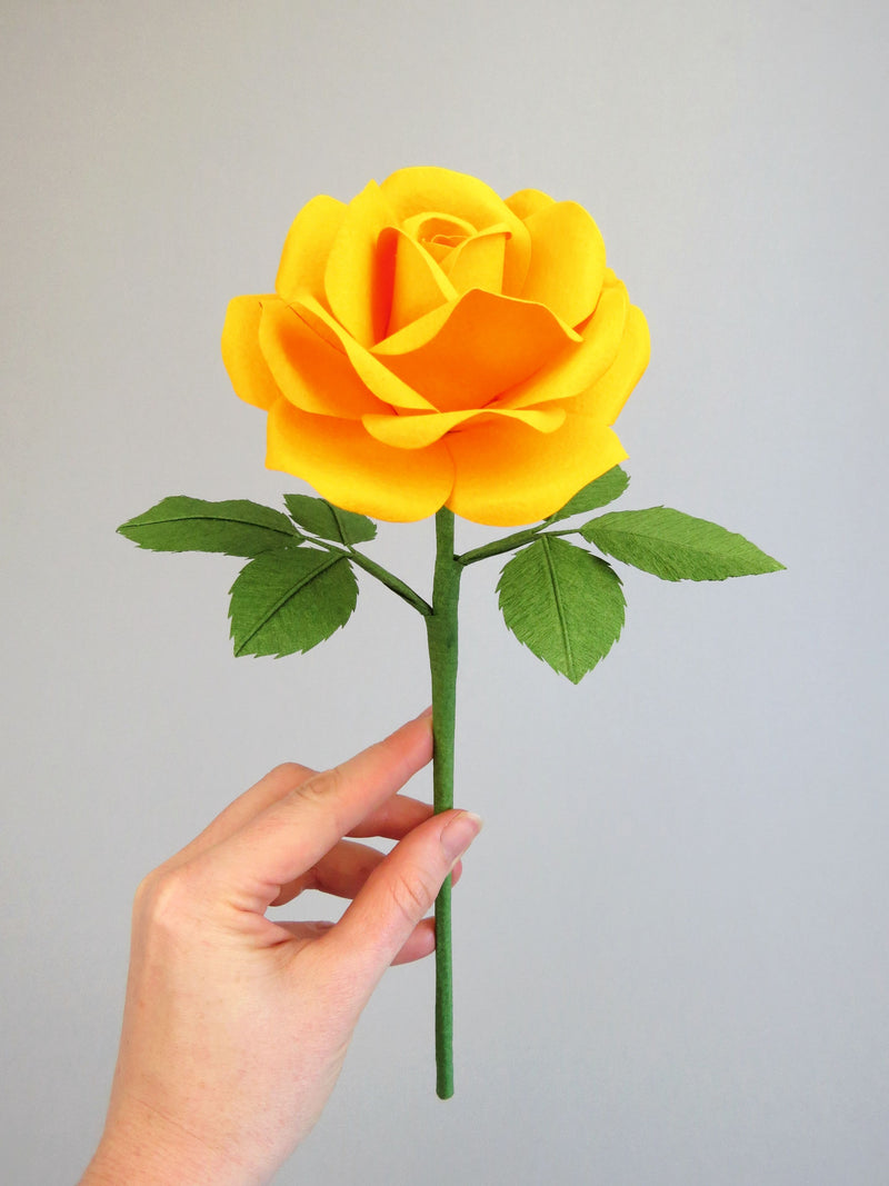 Pale white hand delicately holding the stem of a yellow cotton paper rose with six green leaves