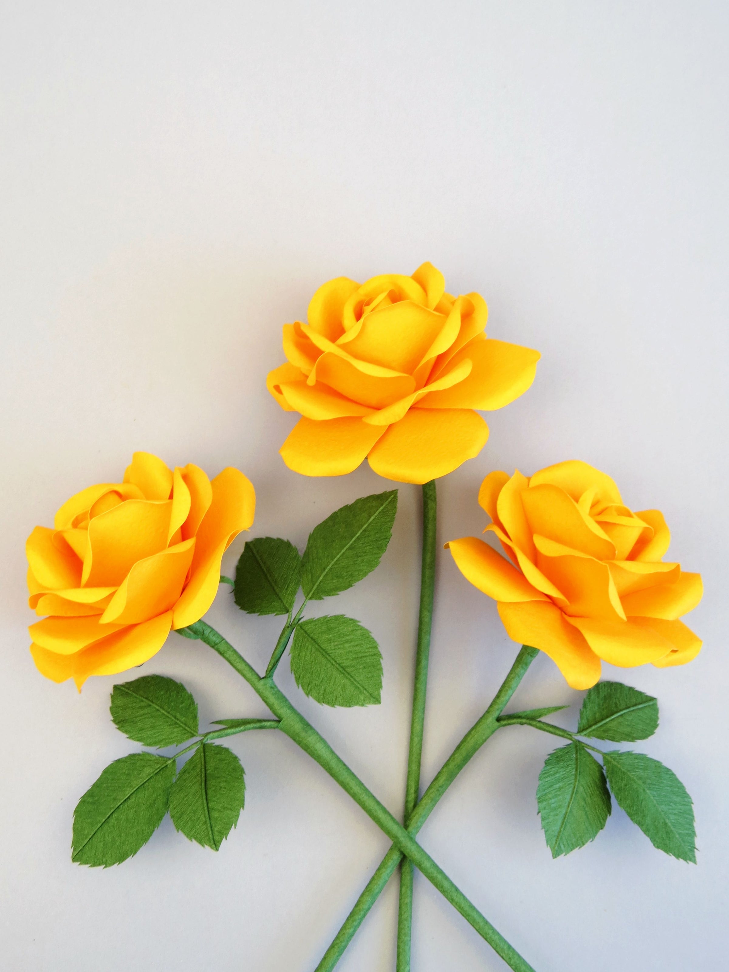 Three yellow cotton paper roses randomly lying next to each other on a grey background. The left rose has six green leaves attached, the middle rose has no leaves and the right rose has three green leaves