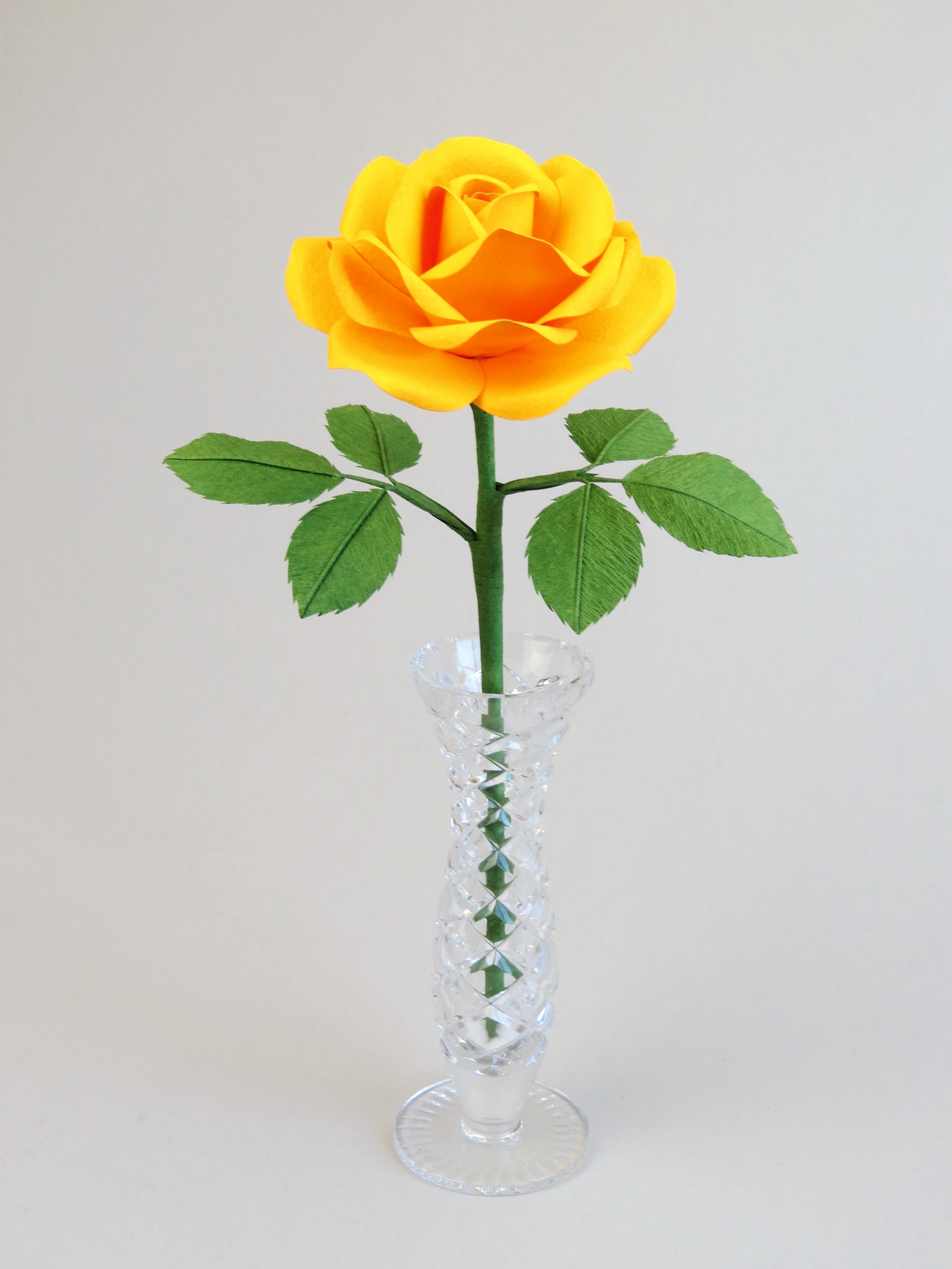Yellow cotton paper rose with six green leaves standing in a narrow glass vase against a light grey background