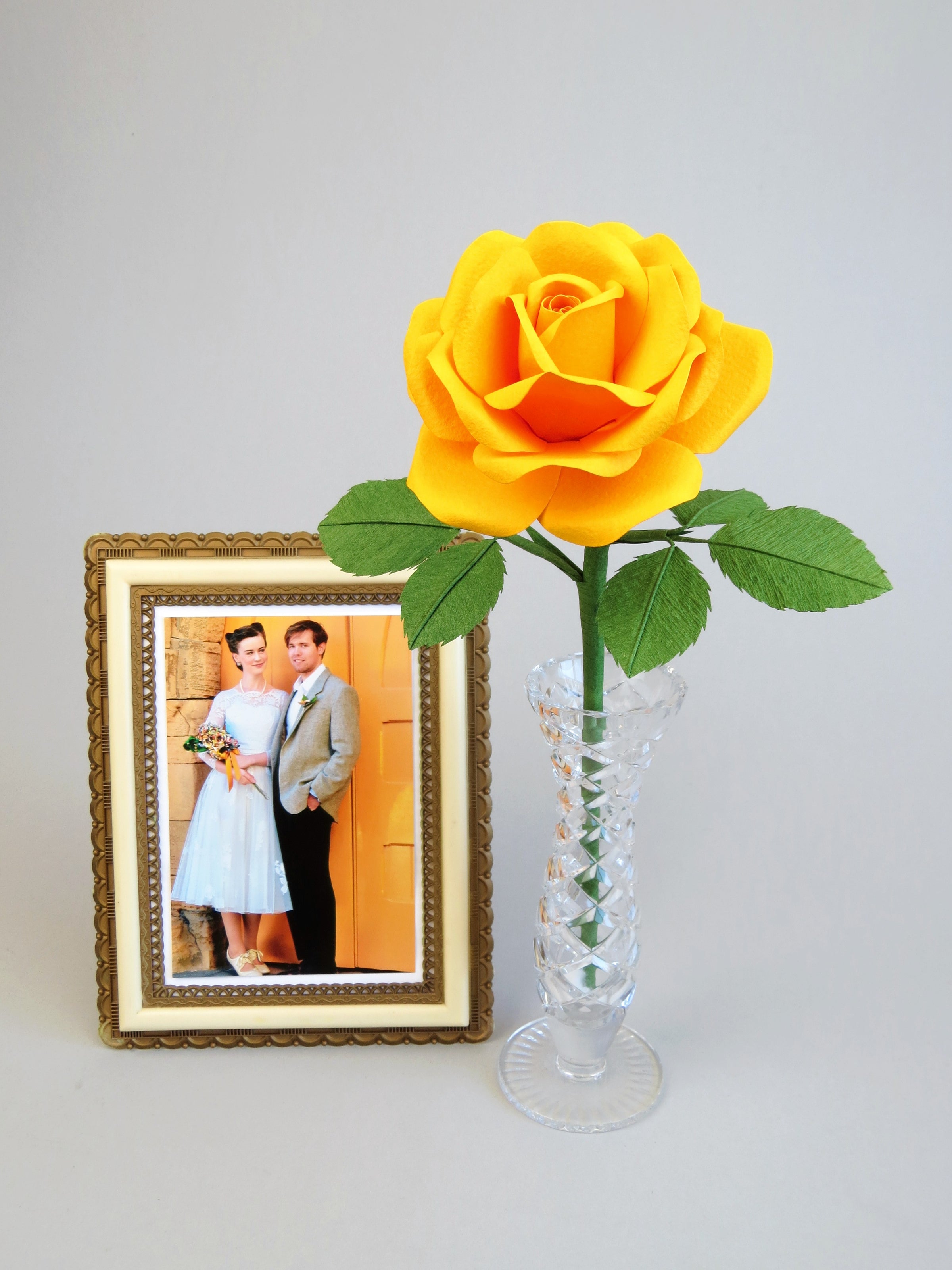 Yellow cotton paper rose with six green leaves standing in a slender glass vase with a framed vintage wedding photo of a happy couple standing beside it against a grey background