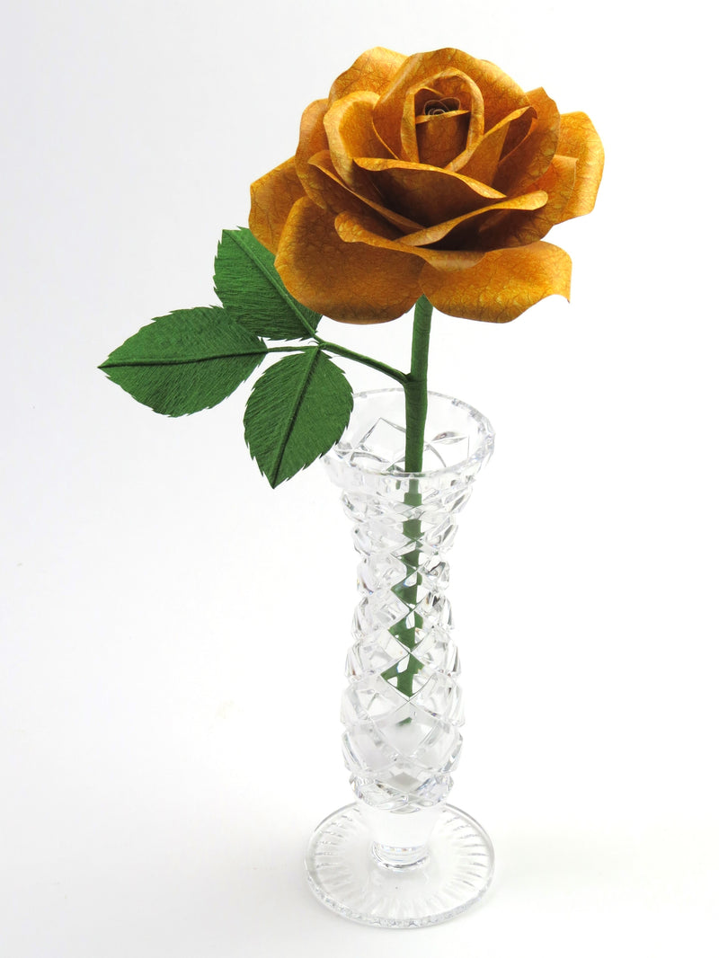 Yellow leather grain paper rose with three leaves standing in a narrow glass vase against a white backdrop