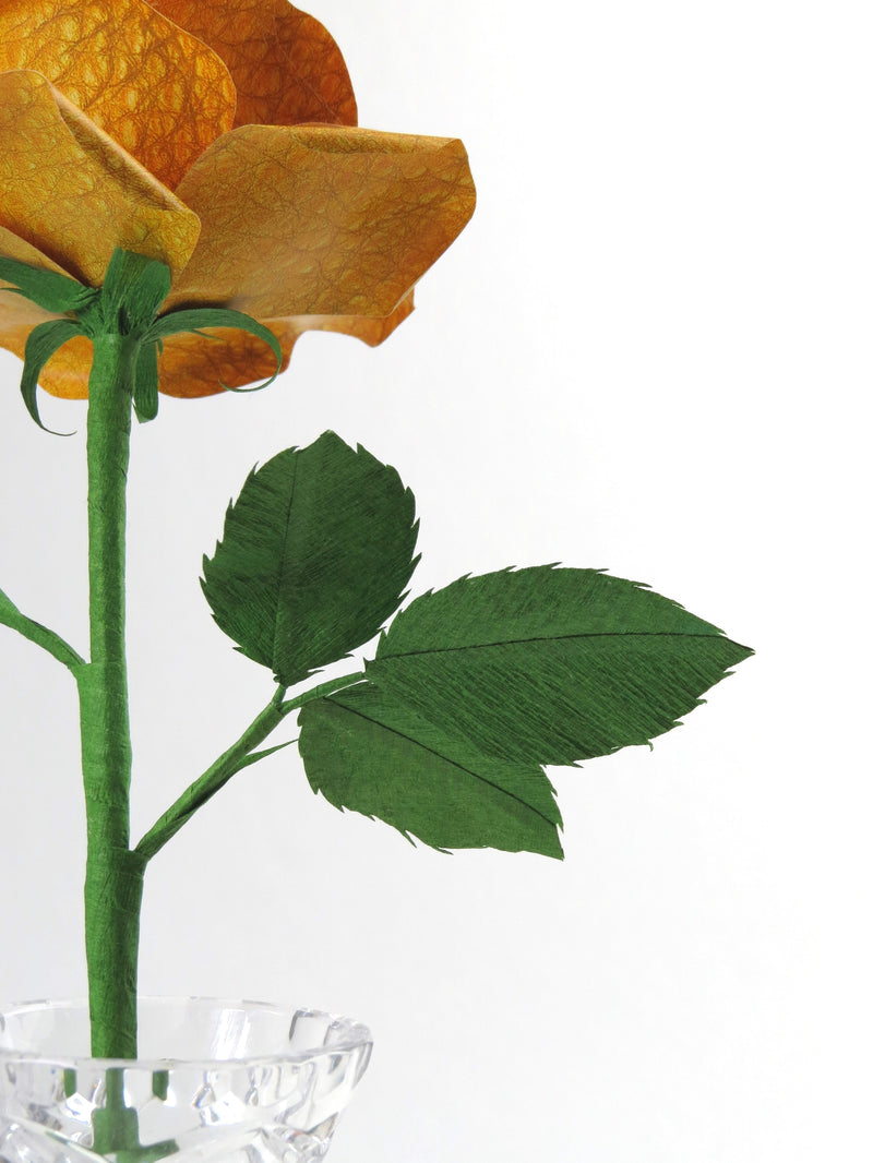 Detailed view of the back of a yellow leather grain paper rose with a green curled calyx underneath the yellow petals, and the green stem, leaves and the tip of the glass vase just in view