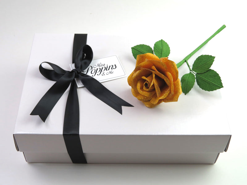 Yellow leather grain paper rose lying on top of a luxury white gift box that has a black satin ribbon tied in a bow with a Miss Poppins and Me gift tag attached
