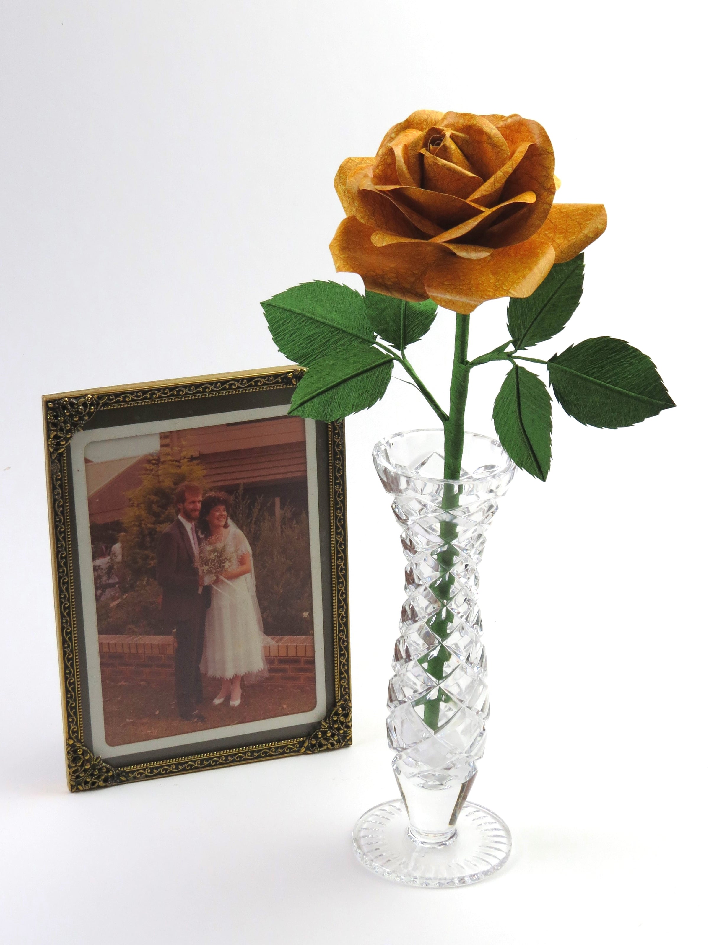 Yellow leather grain paper rose with six green leaves standing in a slender glass vase with a framed wedding photo of a happy couple standing beside it