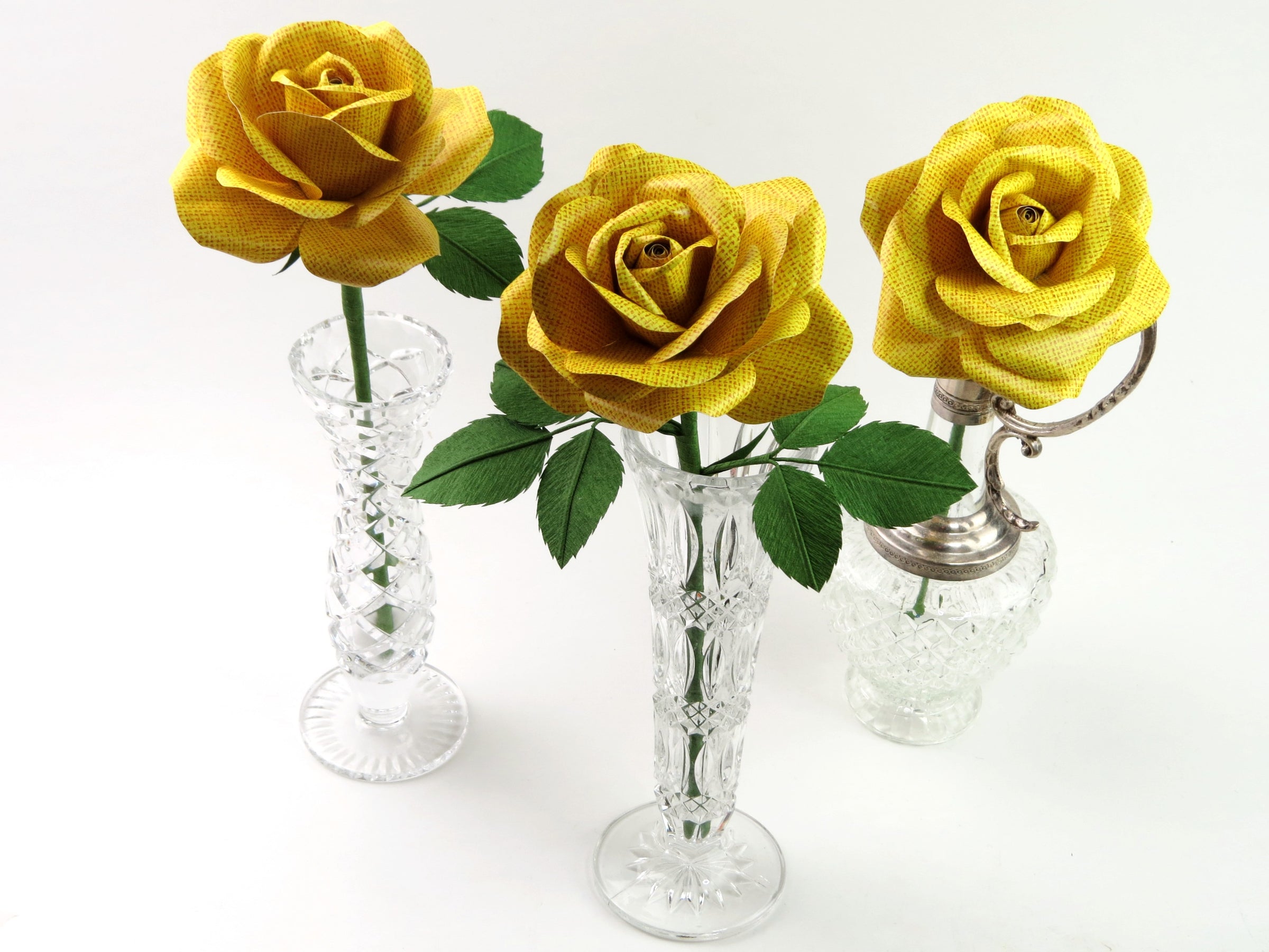 Three yellow linen grain paper roses each standing by itself in three different mismatched glass vases against a light grey background. The left rose has three green leaves attached, the middle rose has six leaves and the right rose has no leaves.