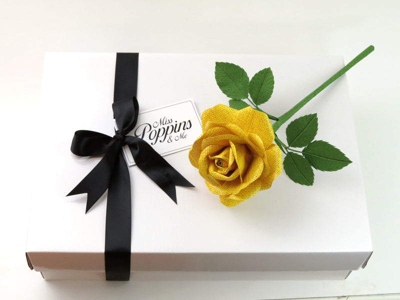 Yellow linen grain paper rose lying on top of a luxury white gift box that has a black satin ribbon tied in a bow with a Miss Poppins and Me gift tag attached