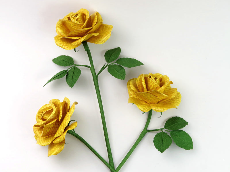 Three yellow linen grain paper roses lying randomly scattered next to each other on a light grey background. The left rose has no leaves attached, the middle rose has six green leaves and the right rose has three leaves. Yellow linen grain paper rose 