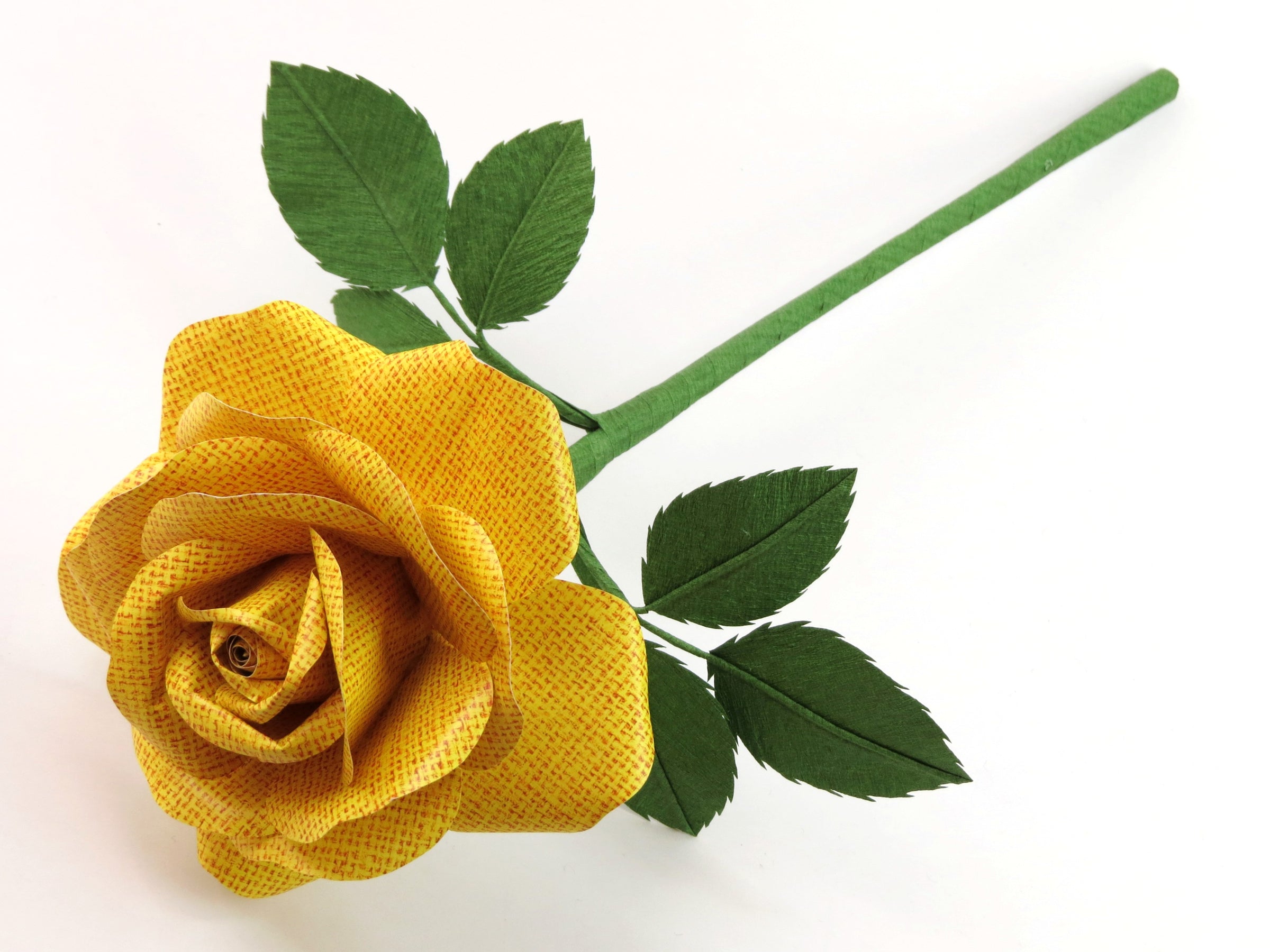 Bright yellow linen grain paper rose lying diagonally across a white background with six green leaves