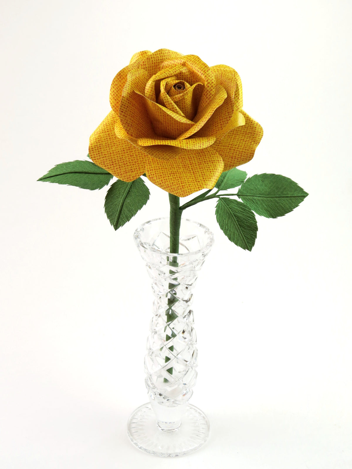 Yellow linen grain paper rose with six leaves standing in a narrow glass vase against a light grey background