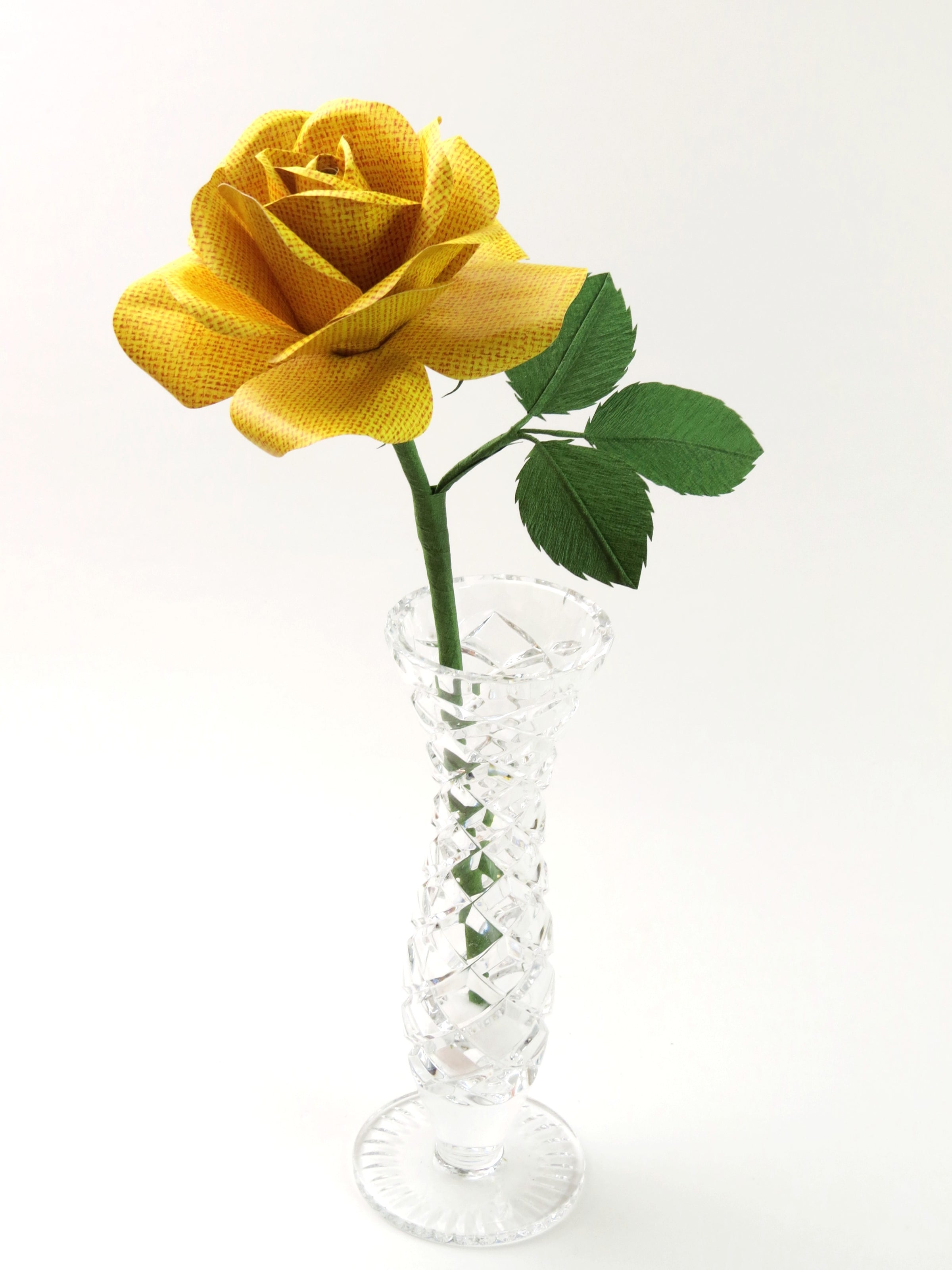 Yellow linen grain paper rose with three leaves standing in a narrow glass vase against a light grey backdrop