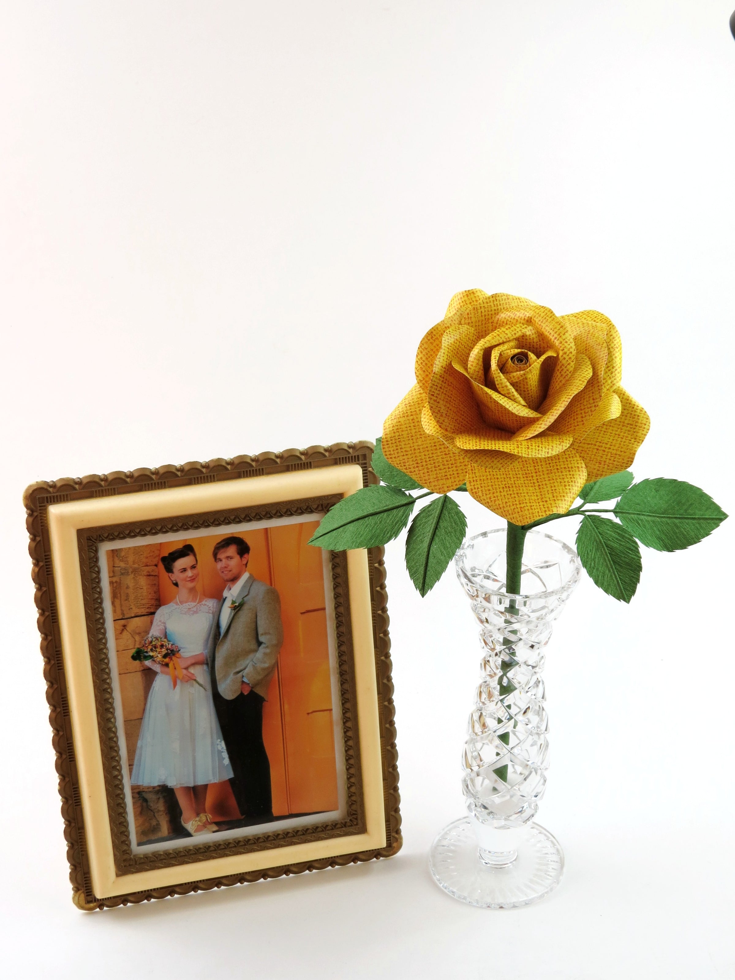 Yellow linen grain paper rose with six green leaves standing in a slender glass vase with a framed vintage inspired wedding photo of a happy couple standing beside it