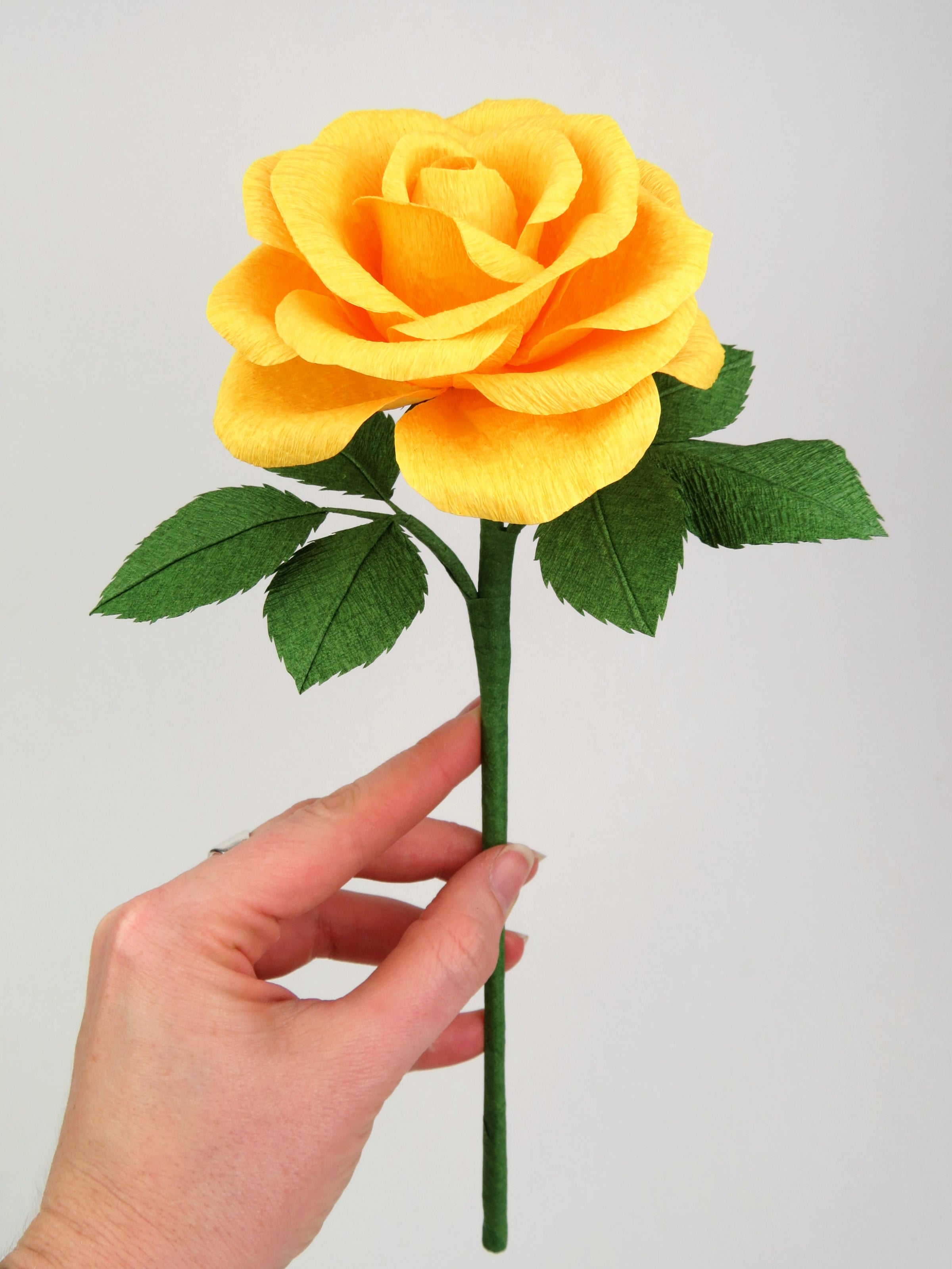 Pale white hand delicately holding the stem of a yellow paper rose with six leaves