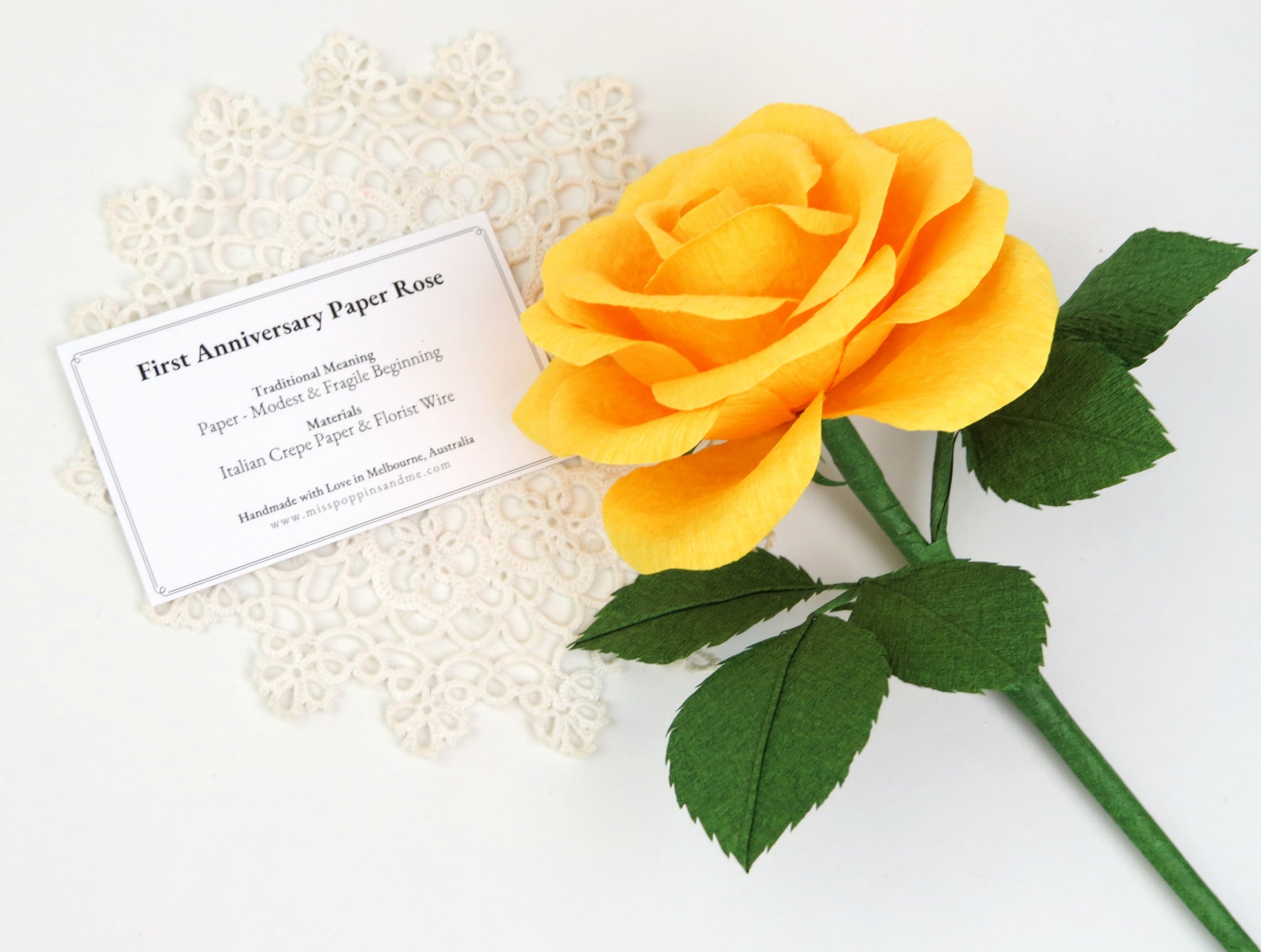 Bright yellow paper rose lying on a small white vintage doily which has a product tag resting on it detailing the meaning and materials of this paper rose