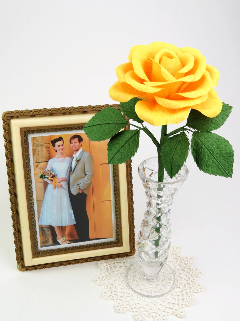 Yellow paper rose with six green leaves standing in a slender glass vase on top of a white vintage doily with a framed wedding photo of a happy couple standing beside it
